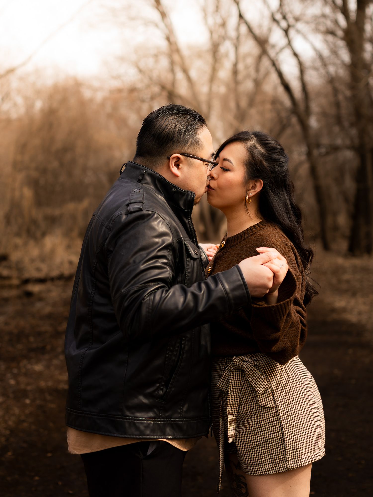 A couple shares an intimate kiss in a natural outdoor setting with bare trees in sepia tones.