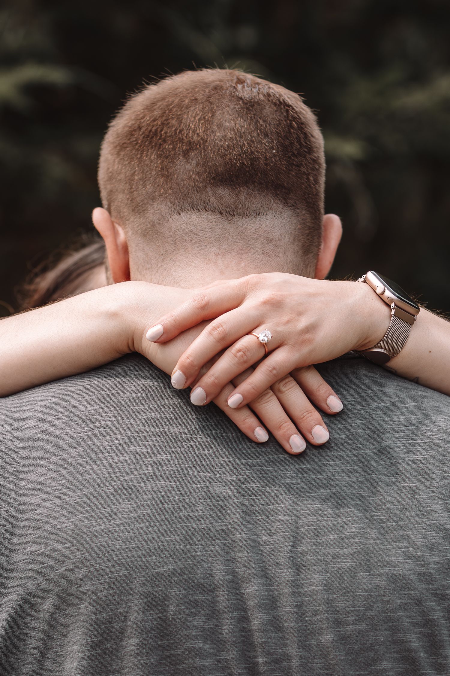 A close-up of hands embracing from behind, showing a diamond engagement ring and wristwatch against a gray shirt.