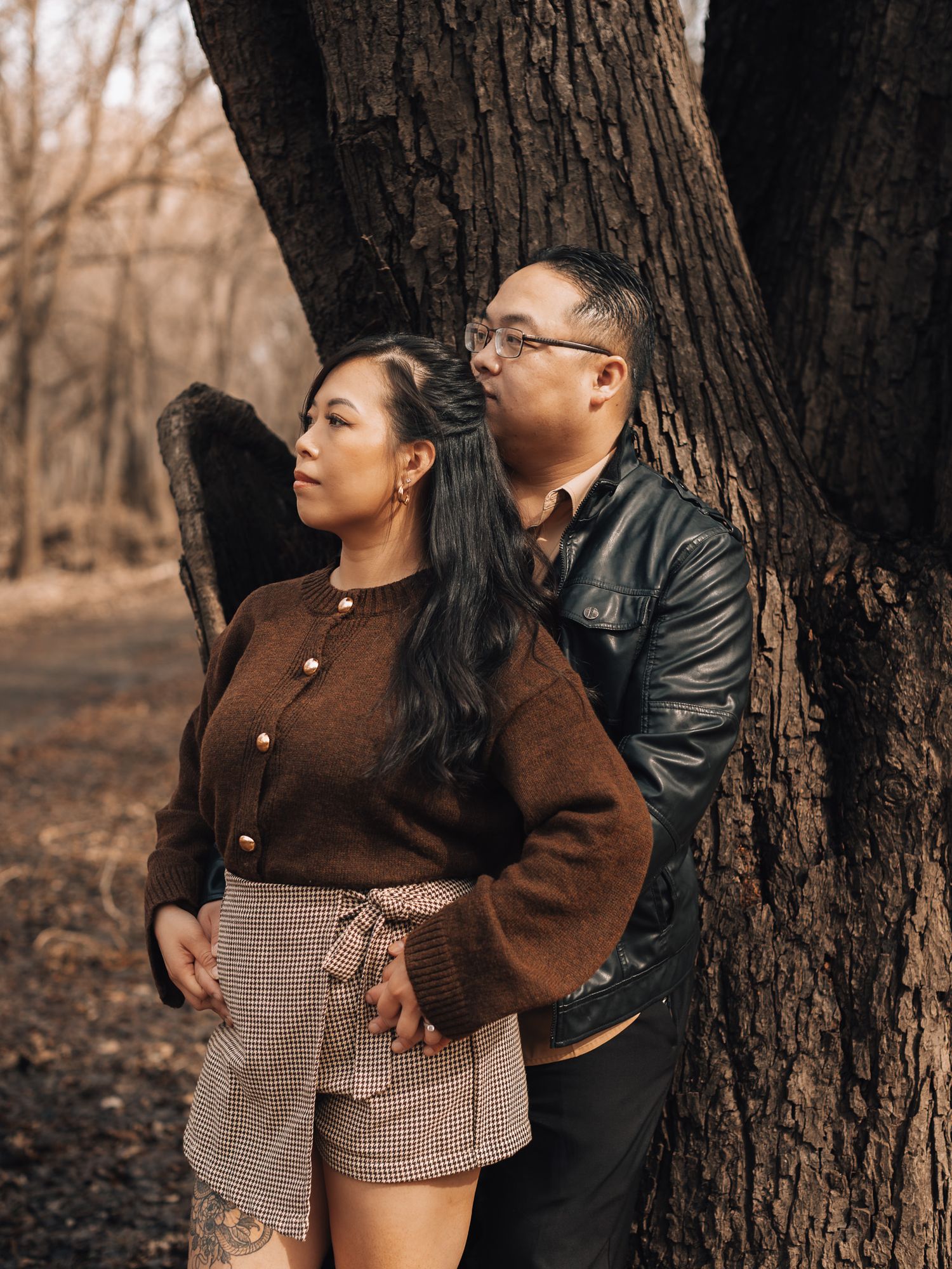 A couple poses intimately against a large tree trunk in an autumn forest setting during a romantic photoshoot.