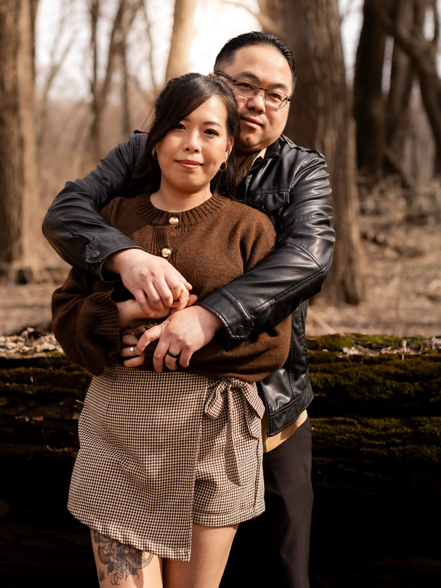 A couple embraces in front of a wooded area during an autumn photoshoot.