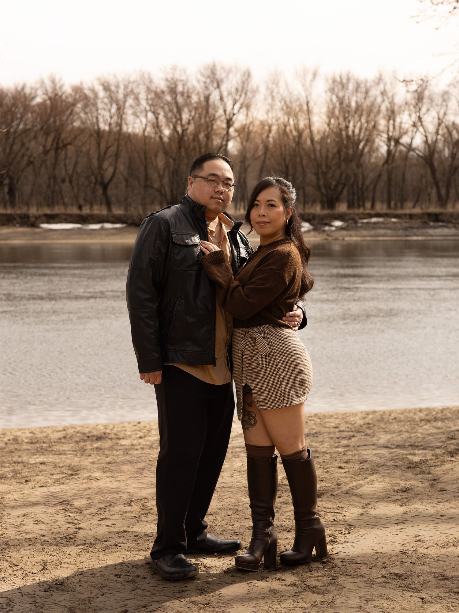 A couple poses together near a lake shore on a winter day with bare trees in the background.