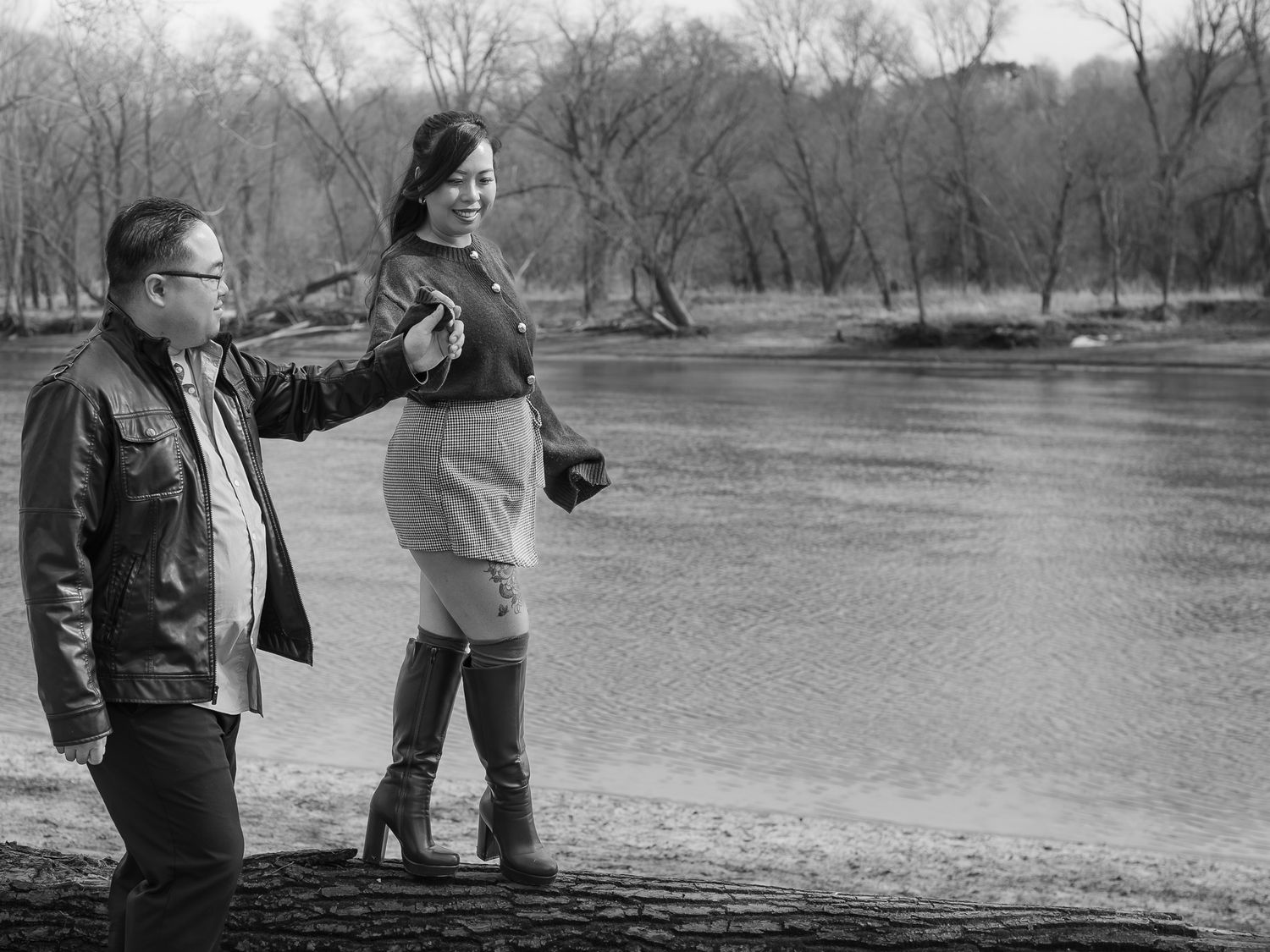 Couple walking together in a winter park landscape with bare trees and a river in the background.
