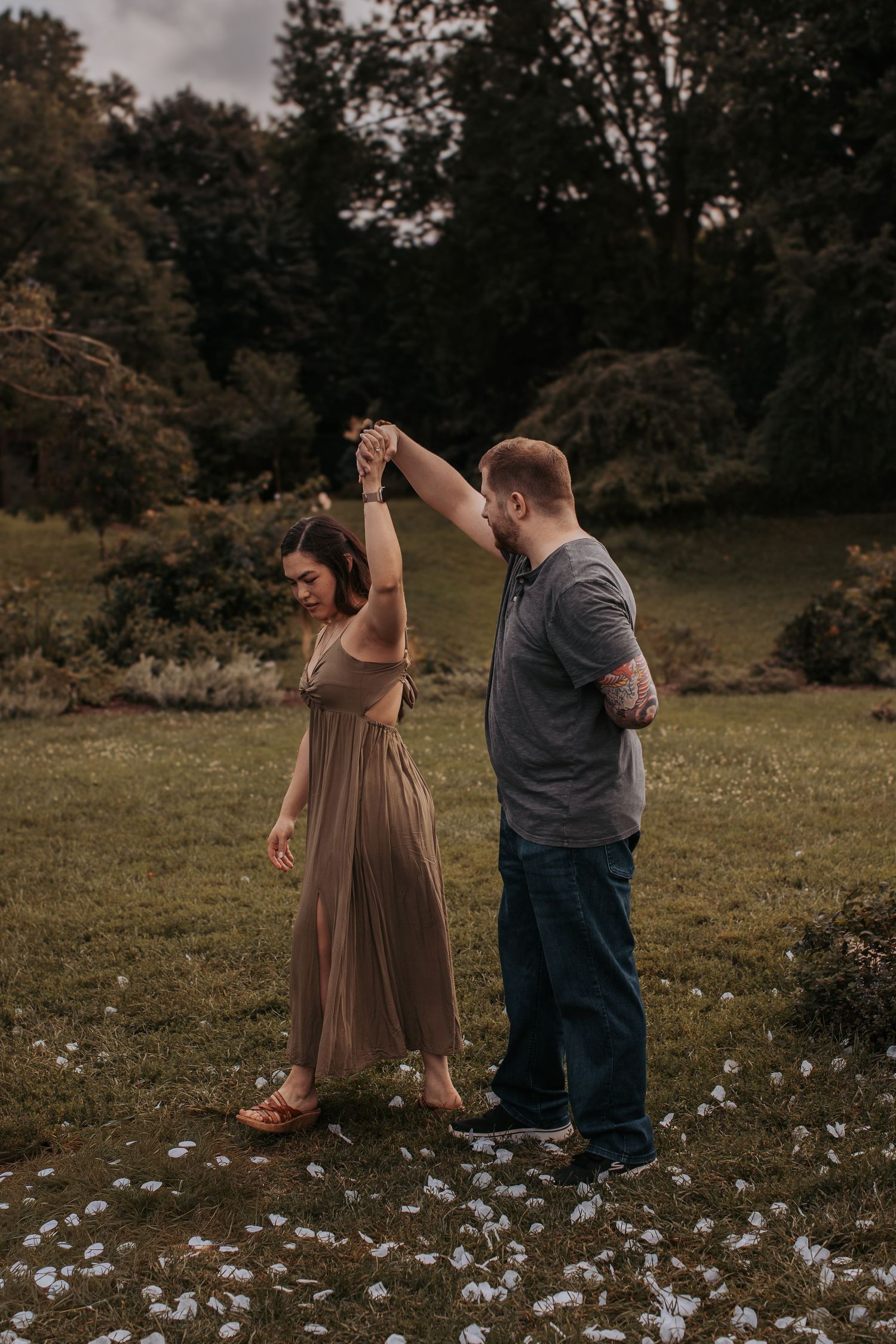 A couple dances together in a meadow surrounded by trees at dusk, with the woman wearing a flowing brown dress.