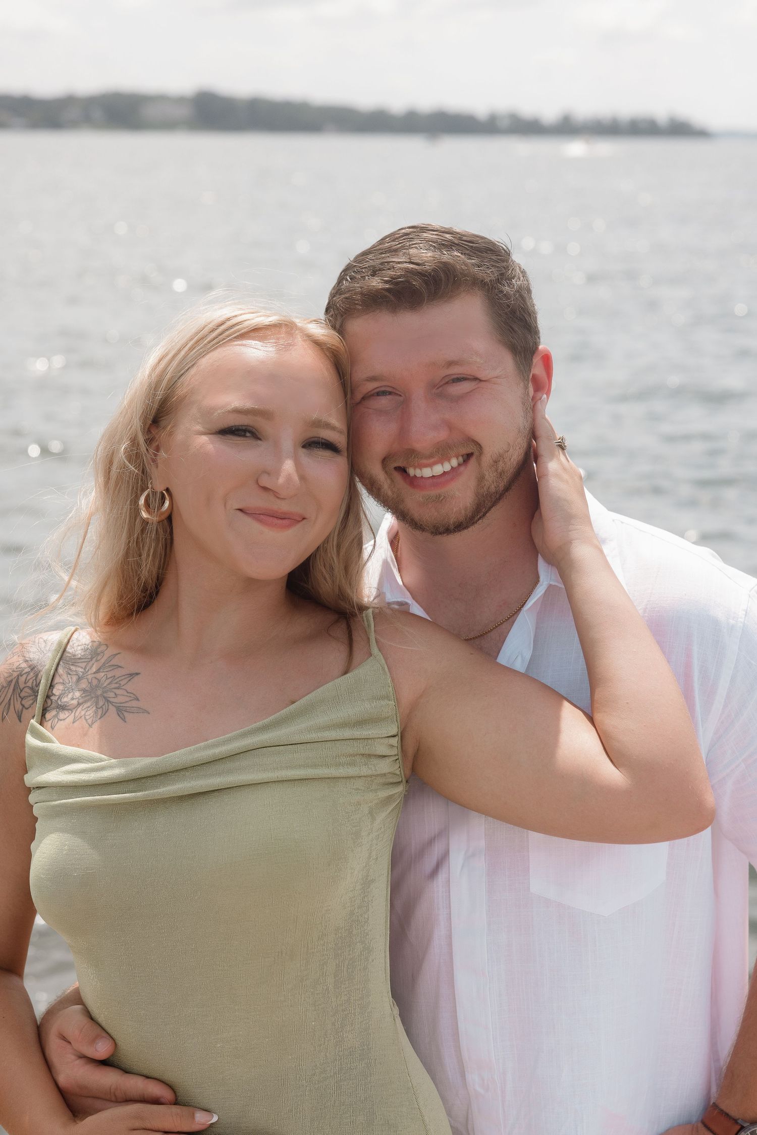 Couple poses affectionately by waterfront on sunny day.