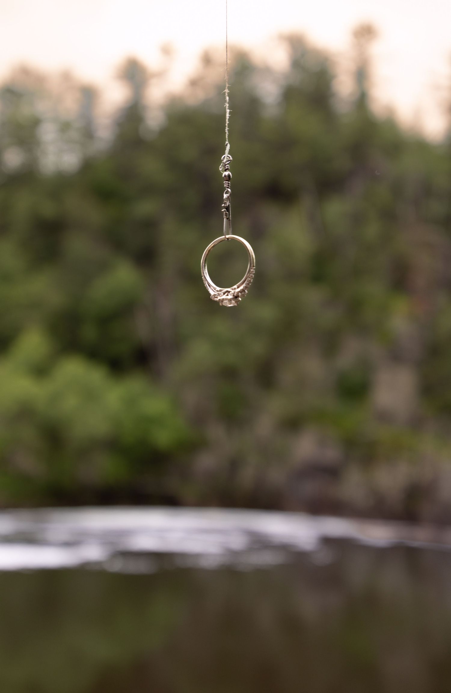 A fishing hook and line hangs suspended in mid-air against a blurred forest and water background.