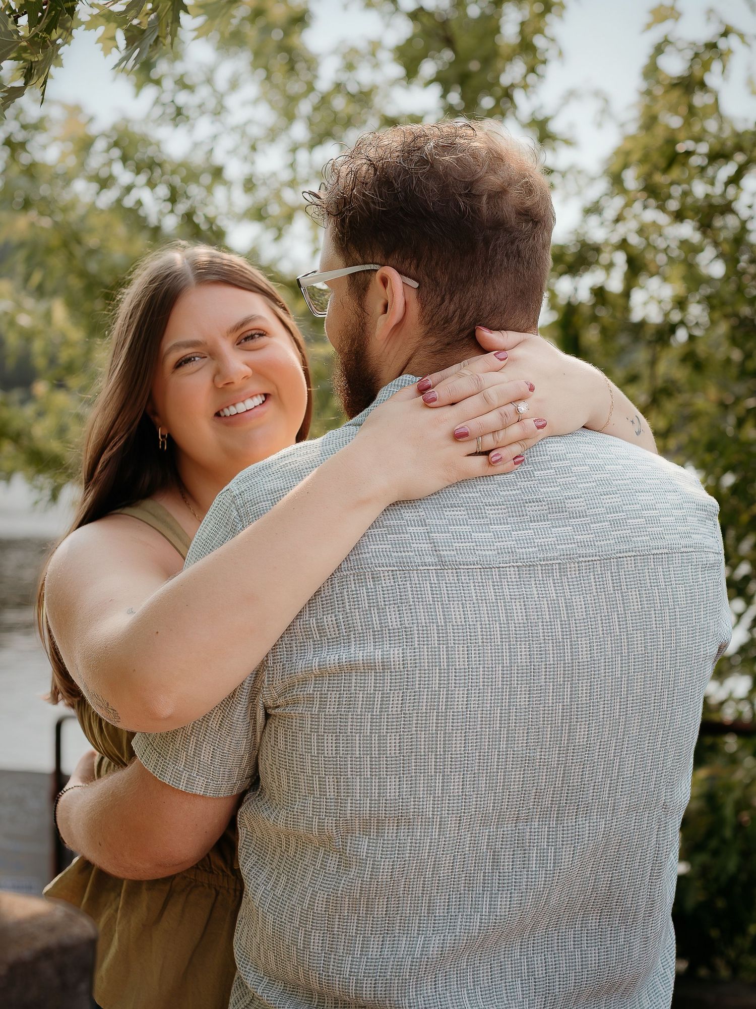 A couple sharing an intimate embrace outdoors with sunlit trees in the background, captured in a candid moment.