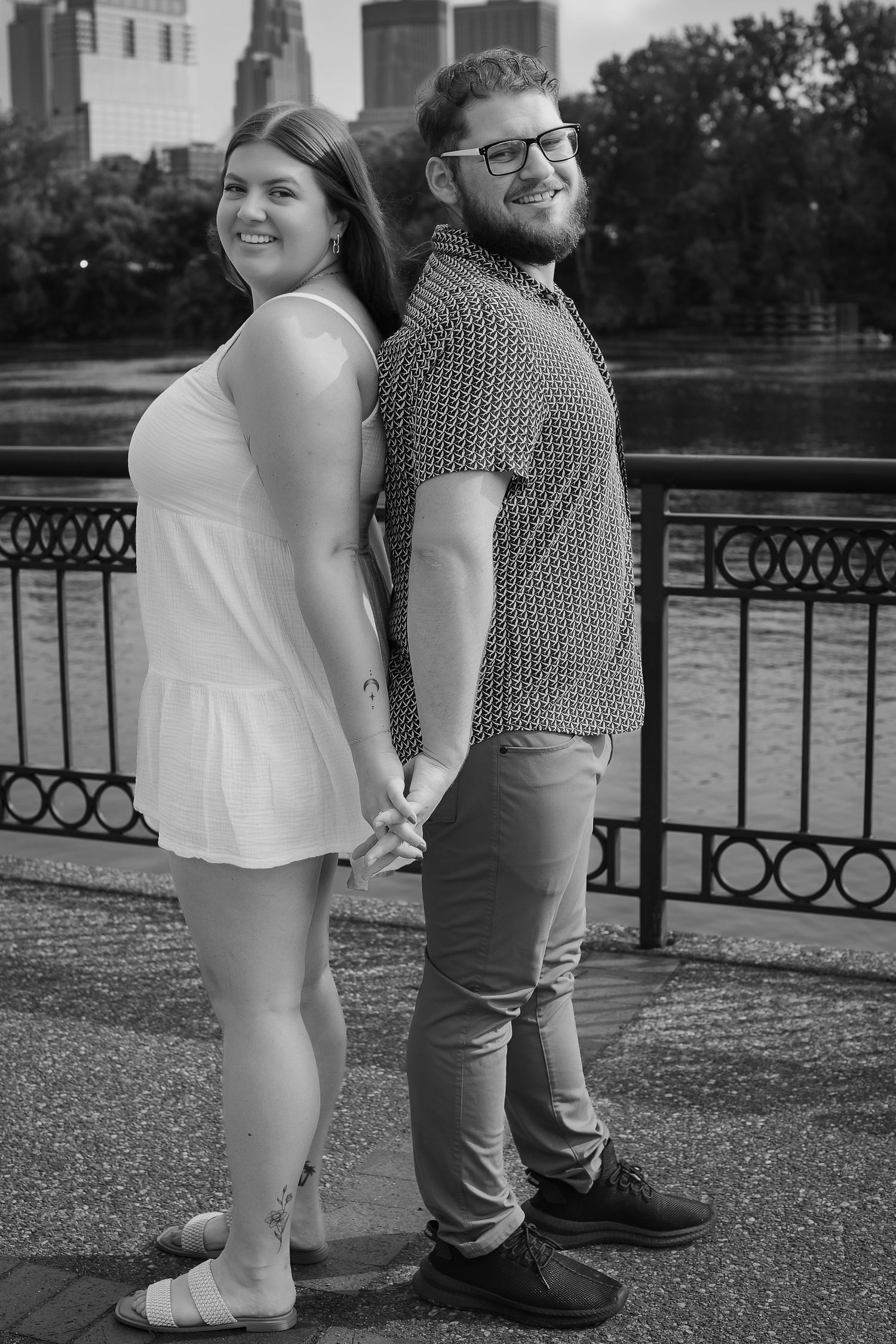 Black and white portrait of a couple holding hands while standing back to back along a railing outdoors.