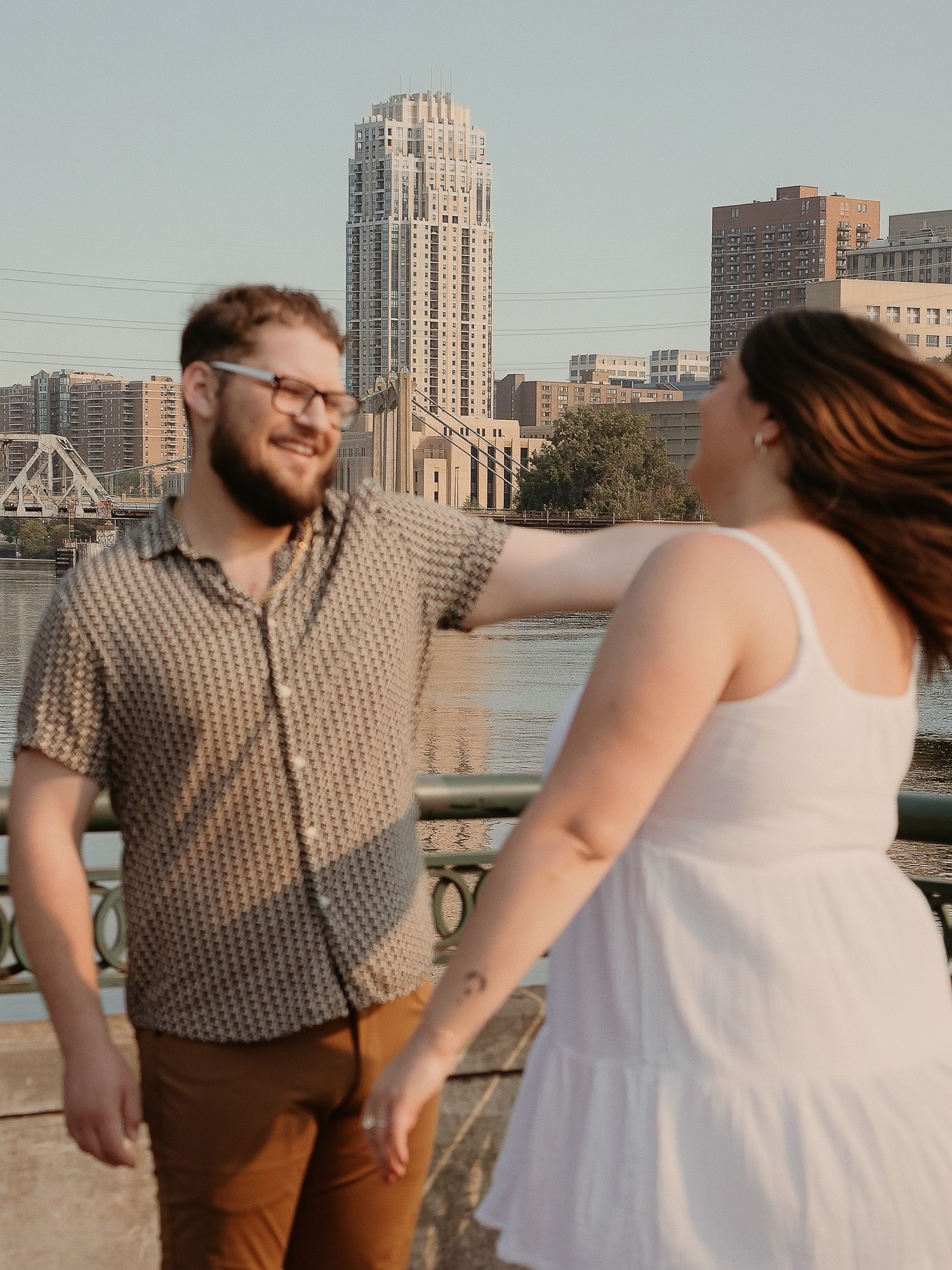 A couple shares a candid moment on a bridge with downtown Minneapolis architecture and buildings in the backdrop.