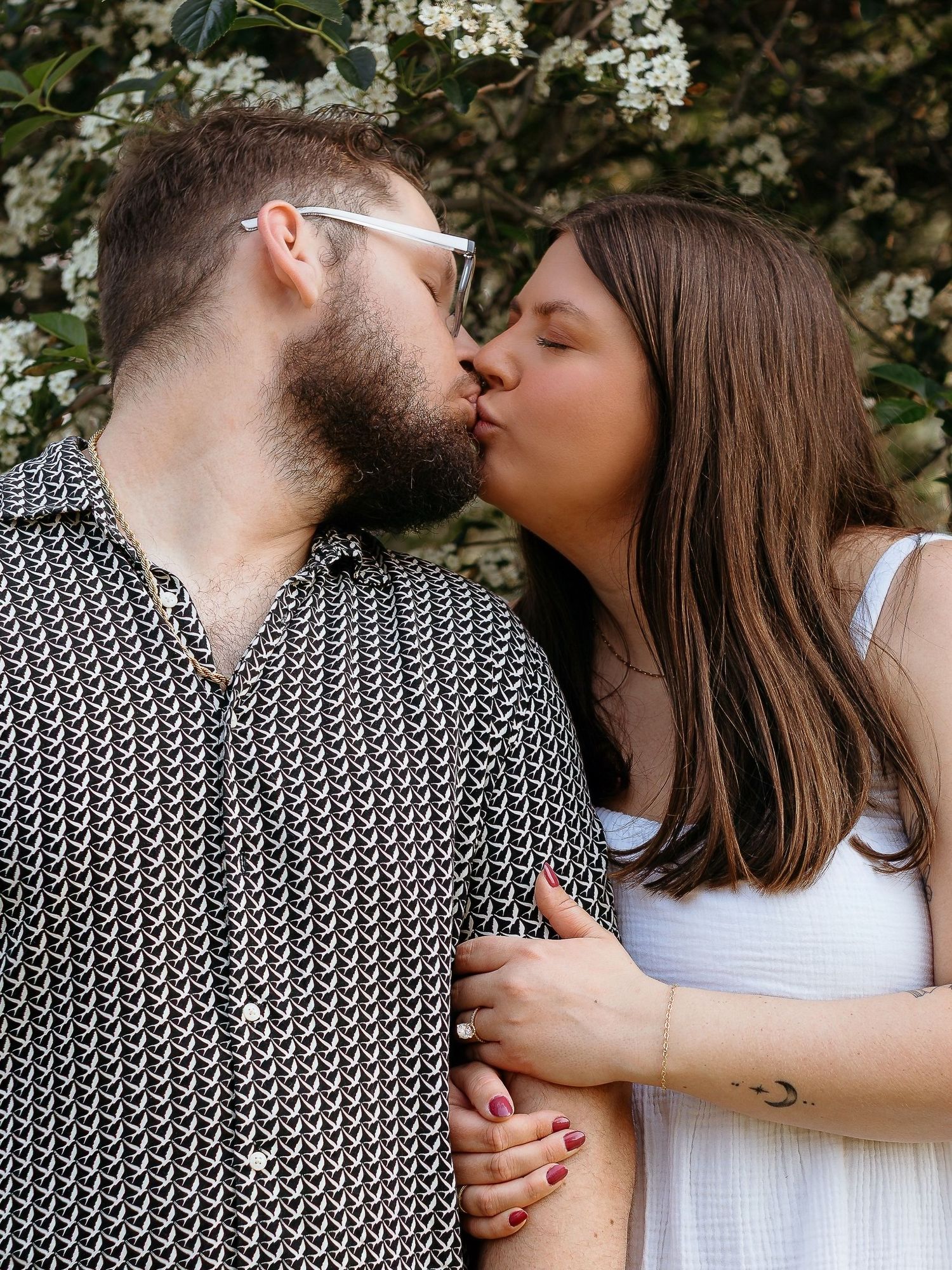 A couple shares a tender kiss while standing against a backdrop of white flowering trees in the springtime.