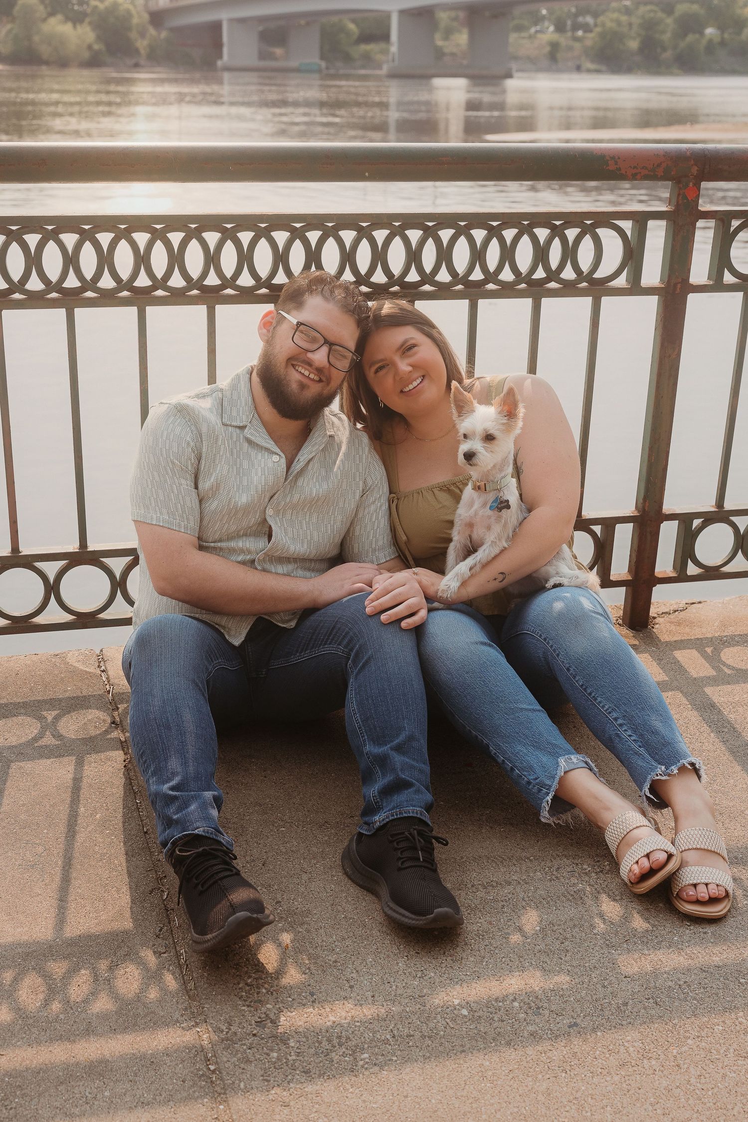 A couple sits together on a railing by the water with their small white dog on a sunny day.