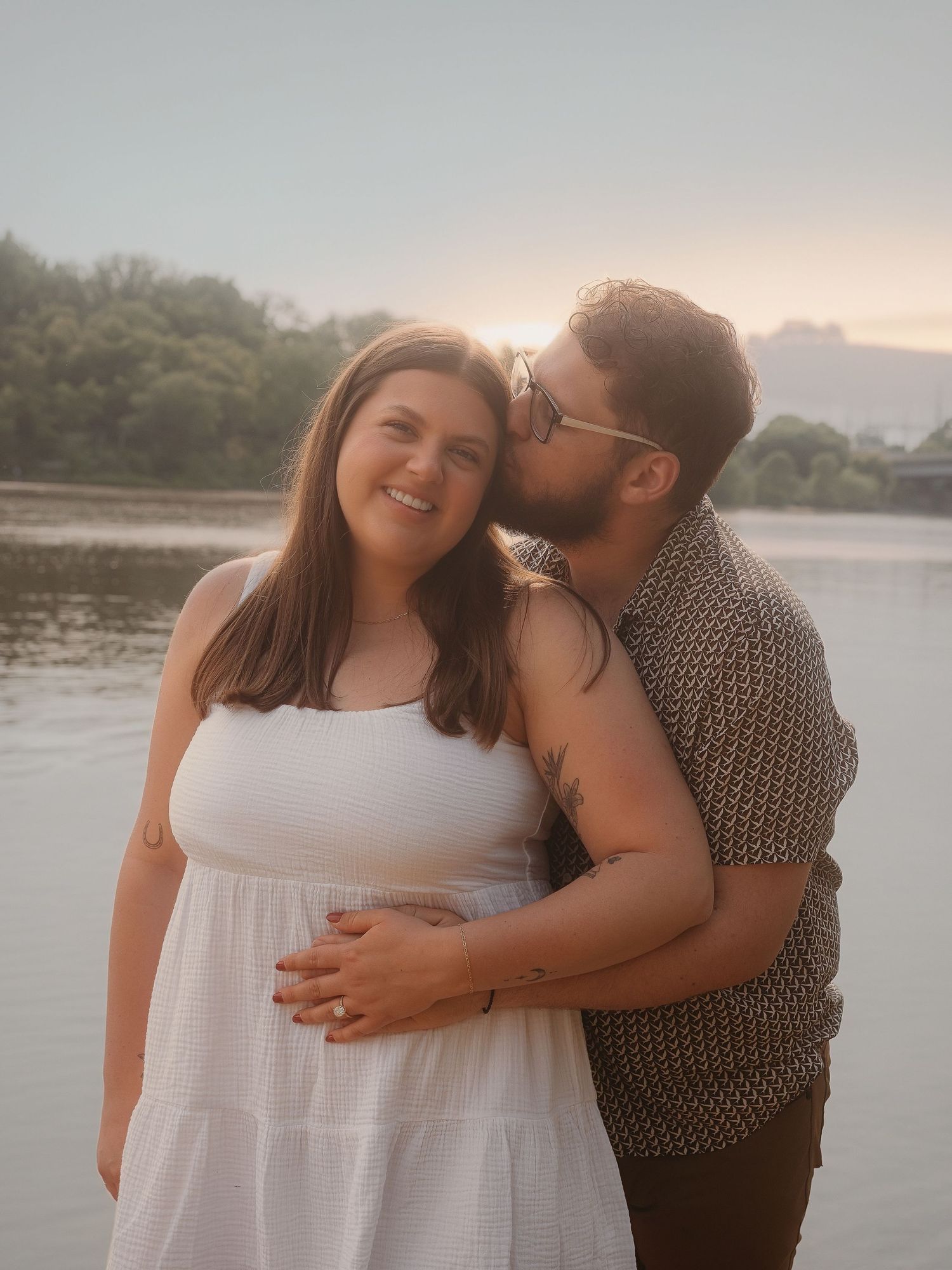A couple embraces during a romantic sunset photoshoot by the water's edge wearing white and patterned clothing.