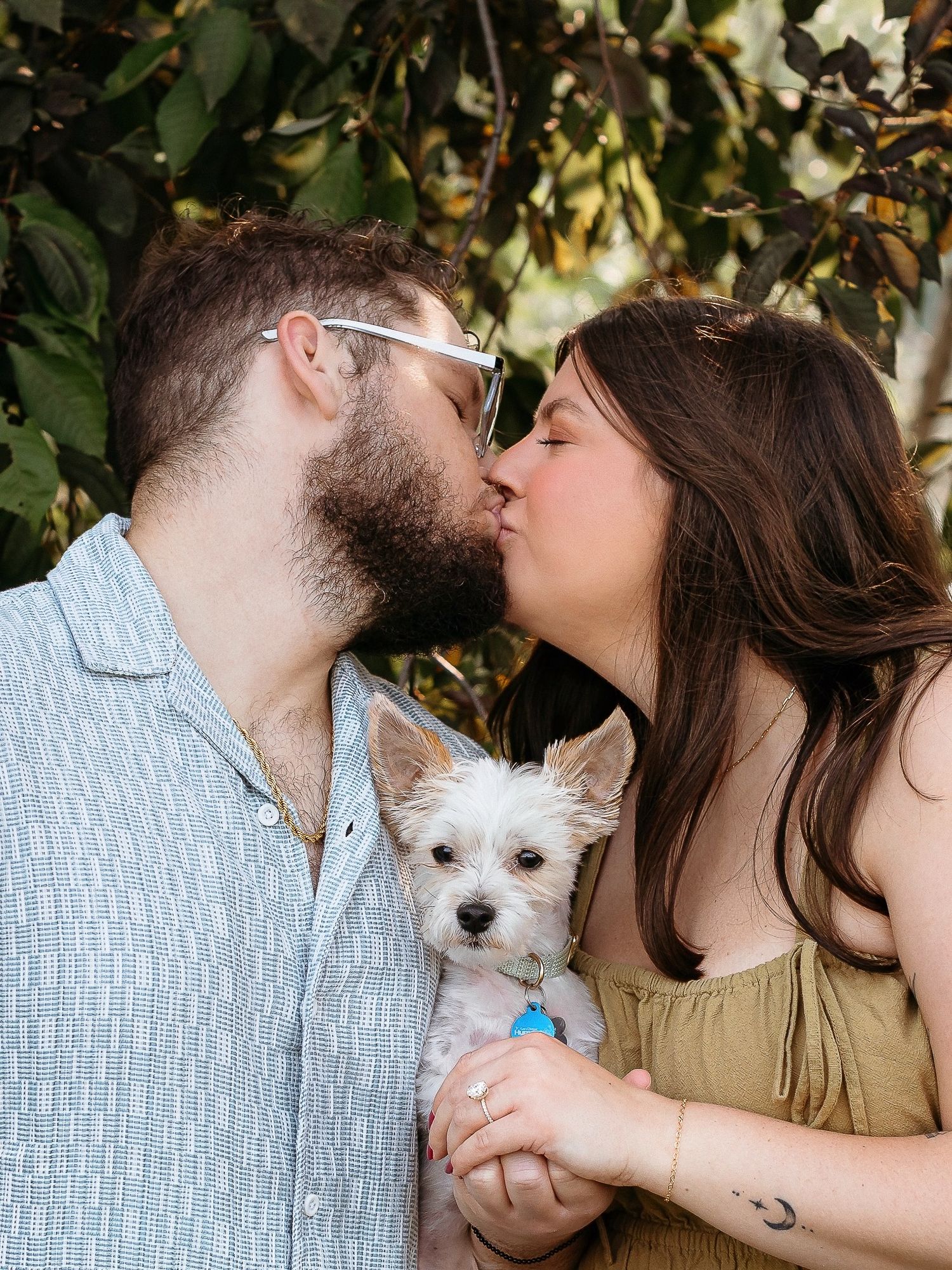 A couple sharing romantic kisses with their small white dog in a series of outdoor photos with green foliage background.