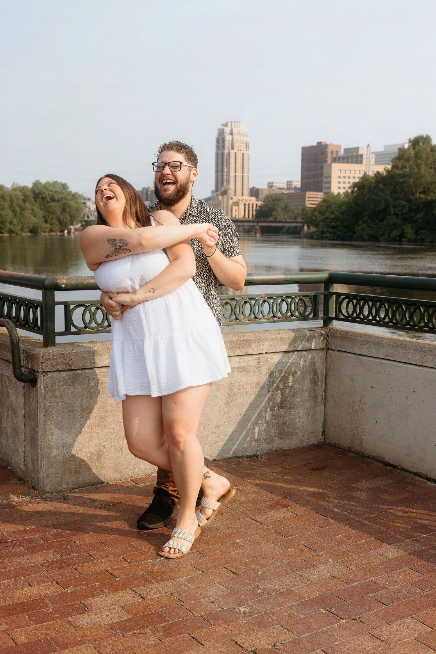 A couple shares a romantic moment dancing on a brick terrace overlooking a river with a cityscape in the background.