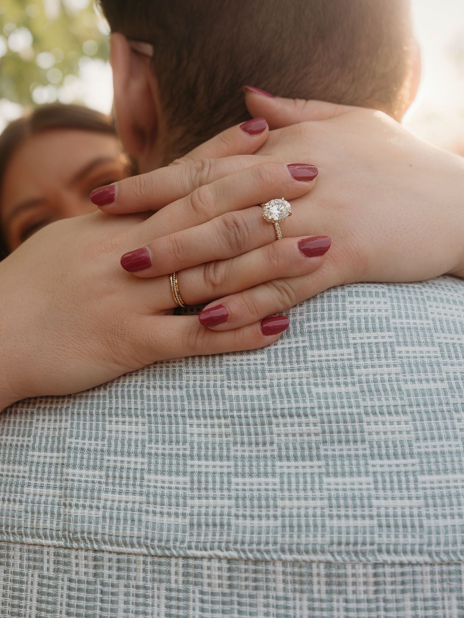 A couple embracing shows a burgundy manicured hand with an engagement ring against a light blue textured fabric background.