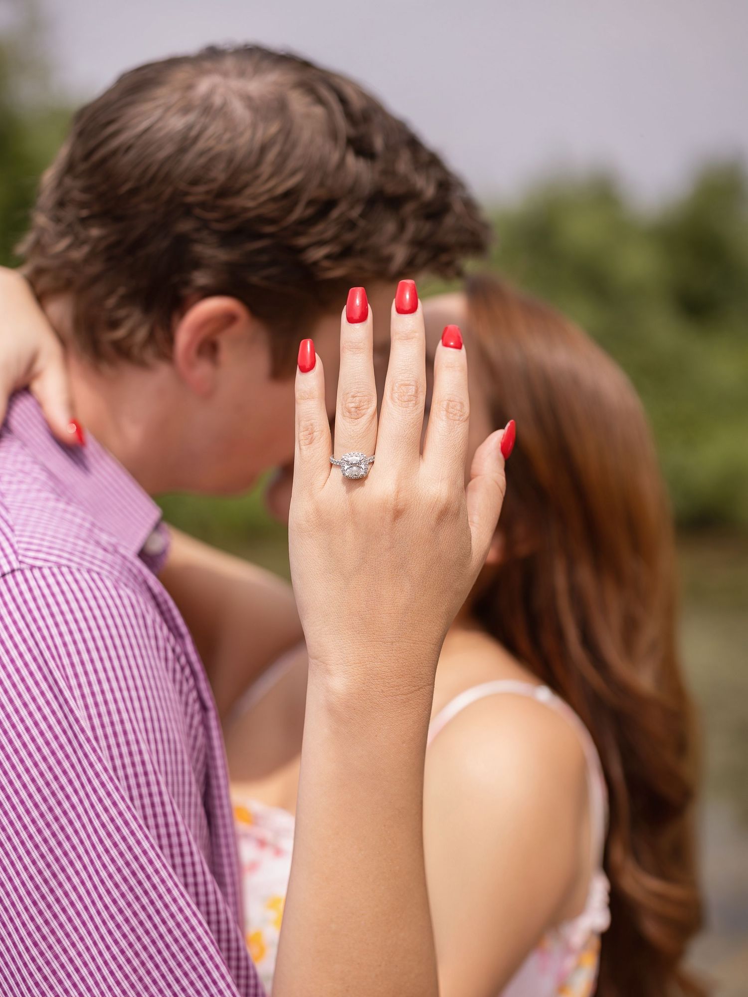 Two hands wearing an engagement ring are shown during an outdoor engagement announcement celebration.