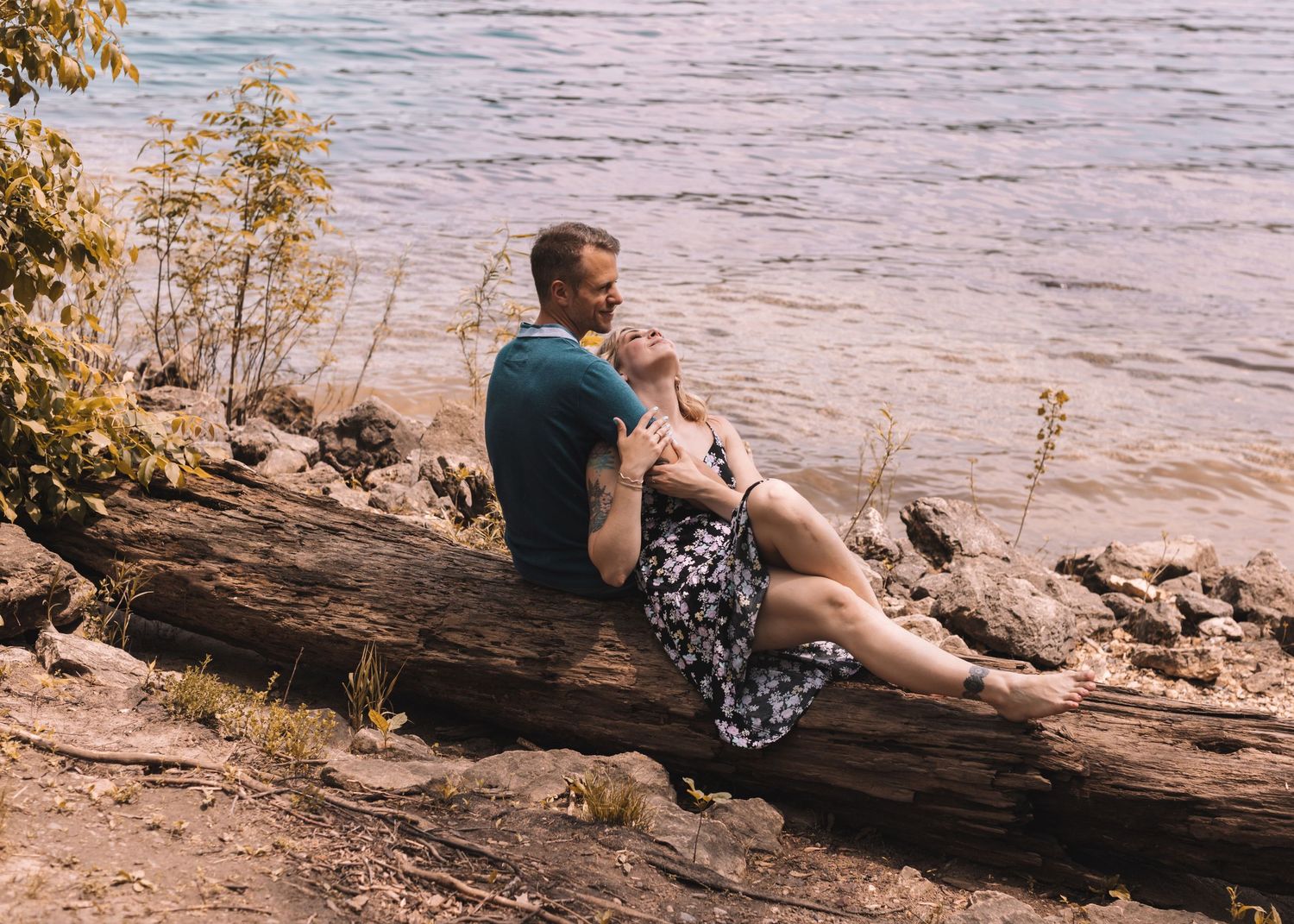 Couple relaxing on fallen log by waterfront during sunset.
