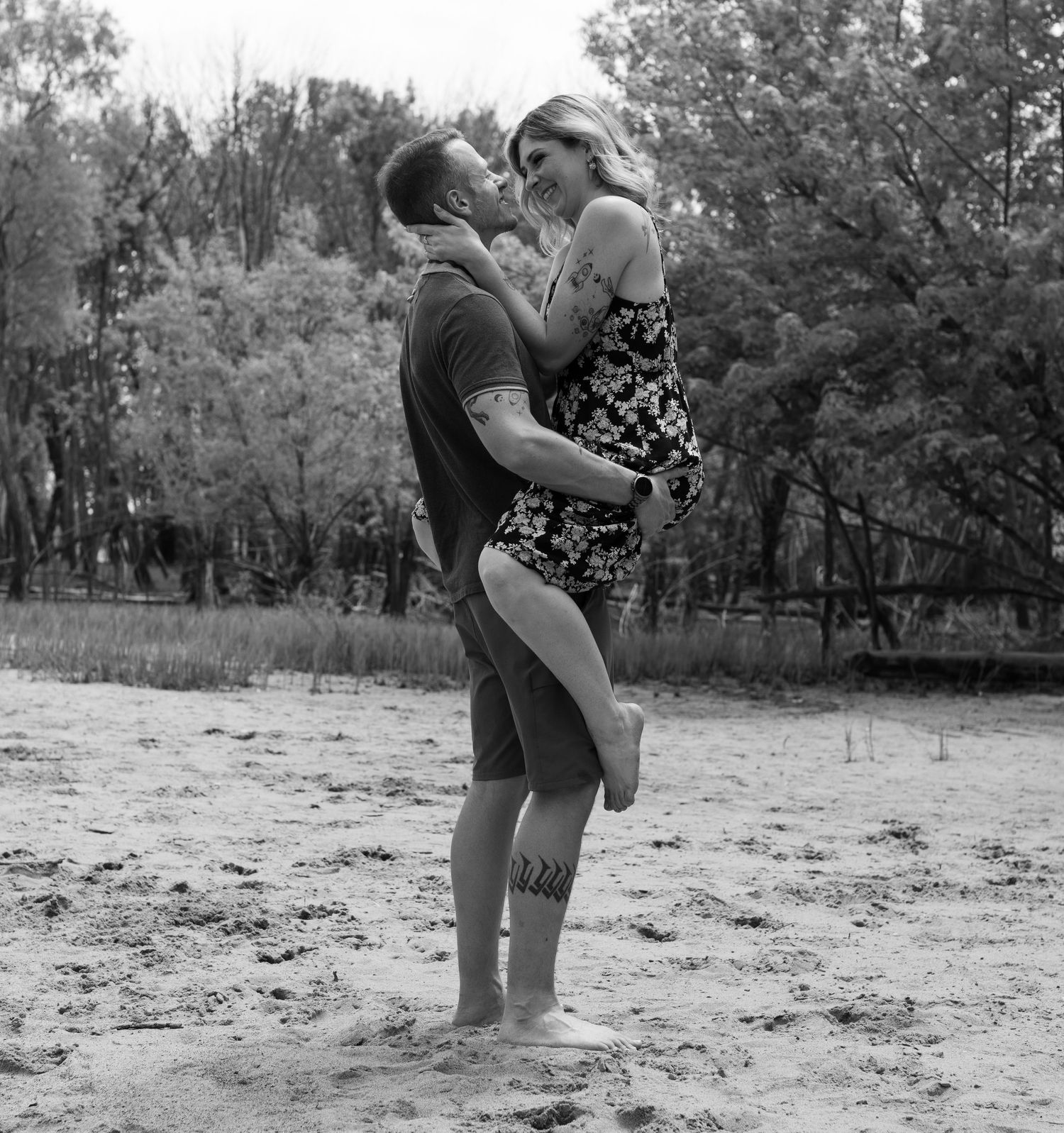 A romantic black and white photo of a couple embracing on a sandy beach with trees in the background.