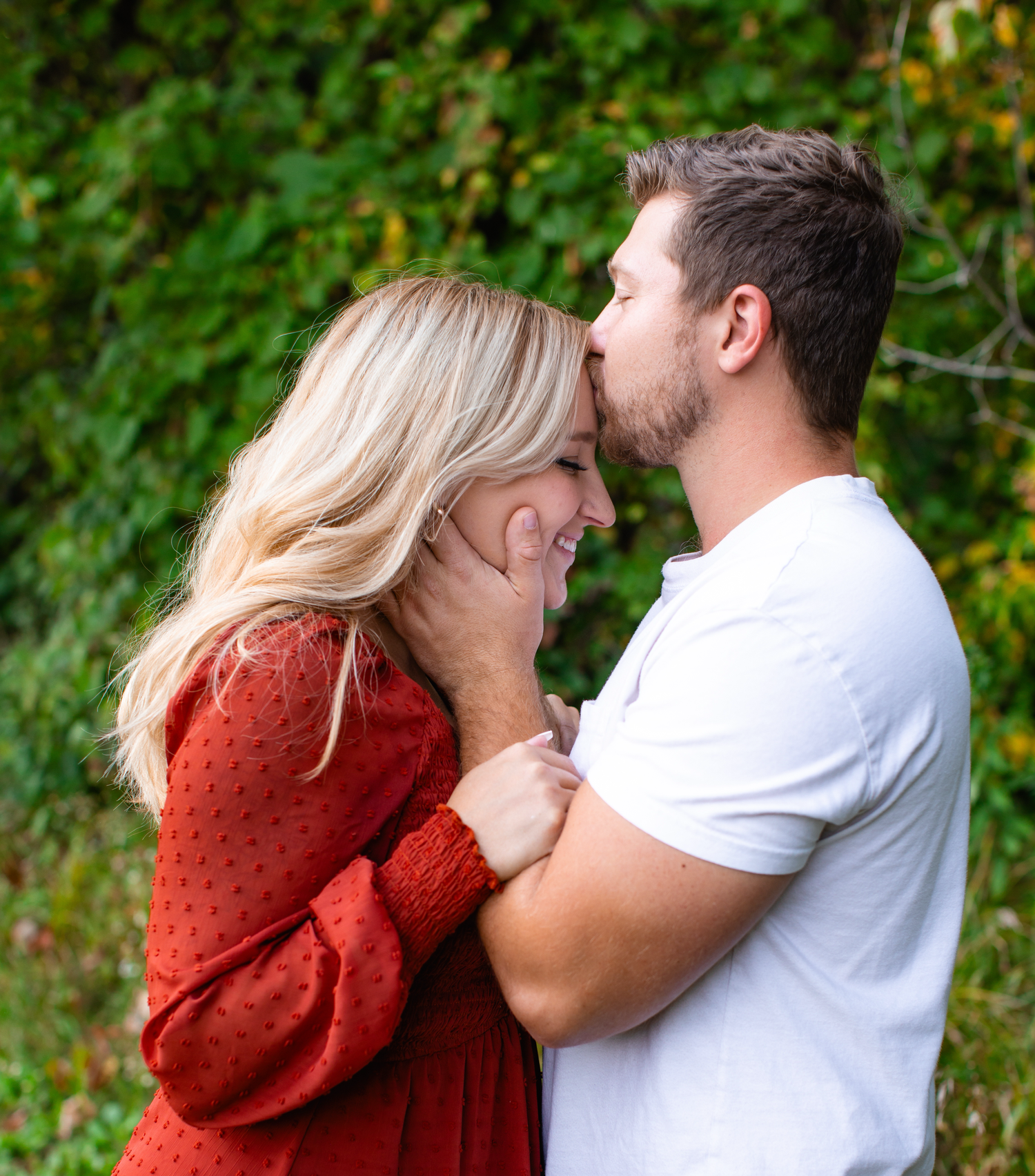 Couple in casual attire sharing tender moment in front of lush green background.