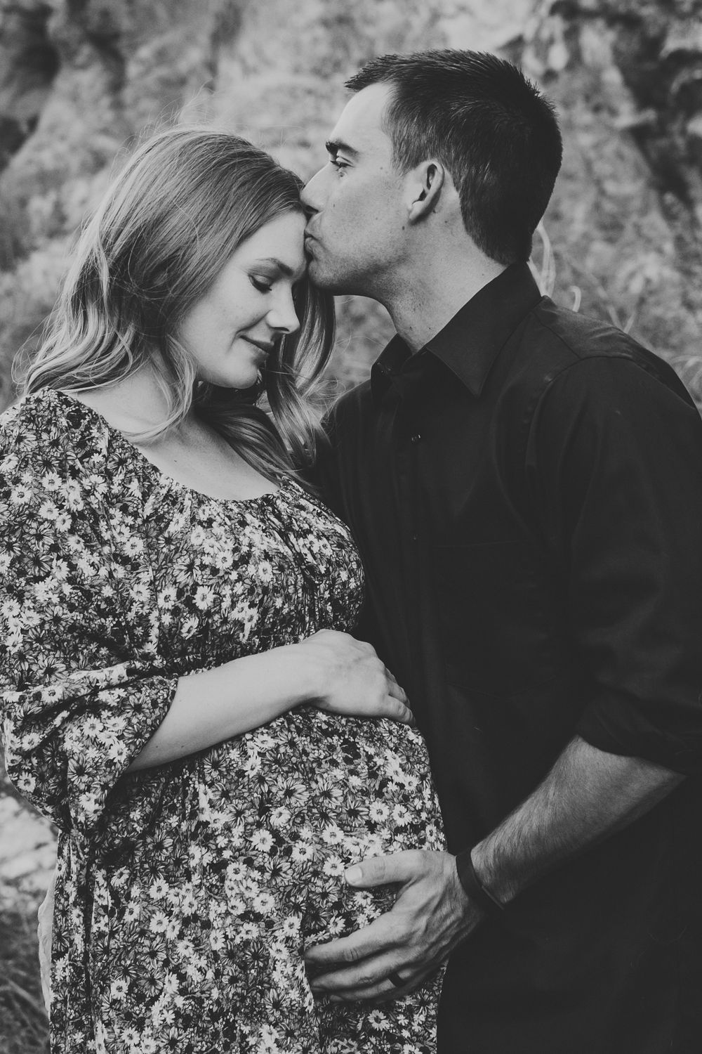 A tender black and white maternity photo shows an intimate moment between a couple outdoors against a rocky backdrop.