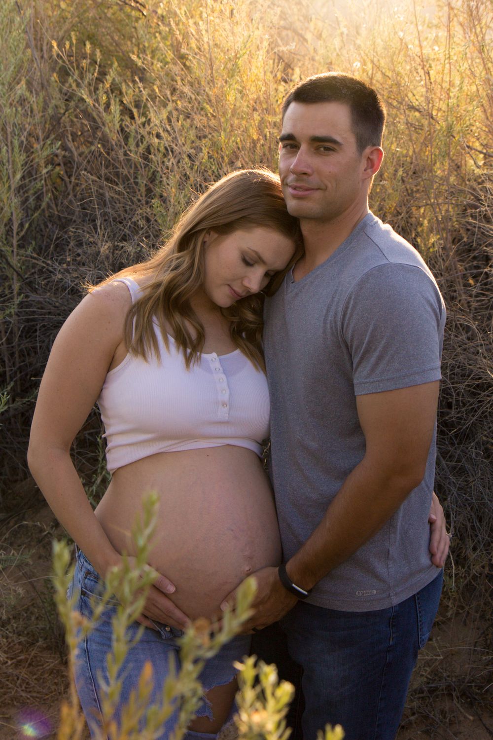 Couple embraces in warm sunset light with yellow wildflowers in foreground during outdoor maternity photo session.