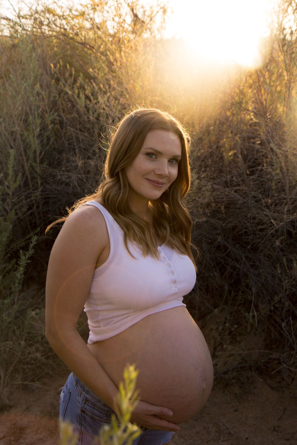 A maternity photoshoot at sunset captures an expectant mother in a white crop top and jeans against golden sunlit foliage.