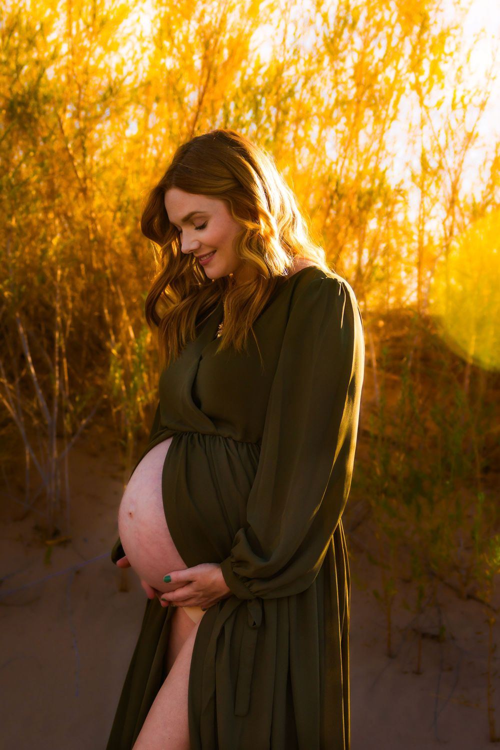 Maternity photo shoot at golden hour shows woman in black dress cradling baby bump against sunlit autumn trees.