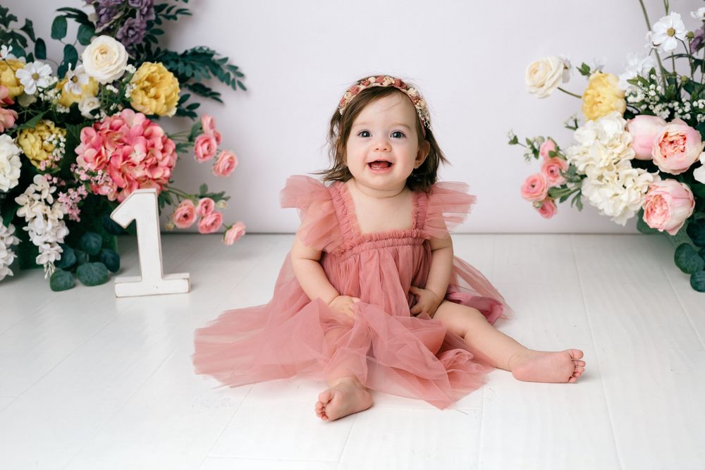 A child in a pink tulle dress poses playfully against a white backdrop decorated with pink and yellow floral arrangements.