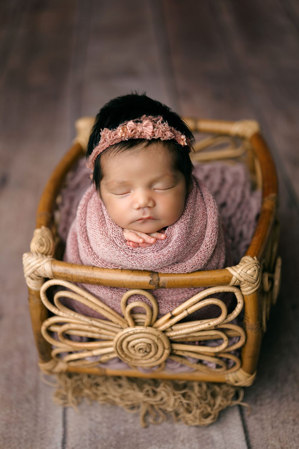 A sleeping newborn swaddled in pink blanket rests peacefully in a decorative woven basket.
