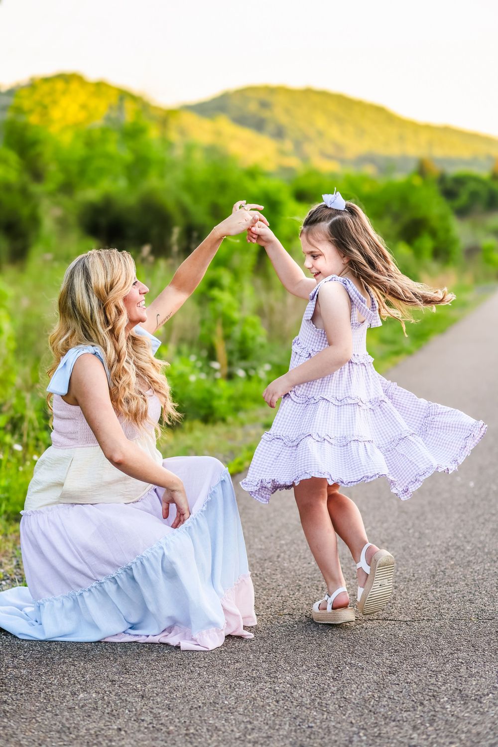 A lifestyle picture of a mother twirling her young daughter in a dress with mountains in the background at golden hour sunset.