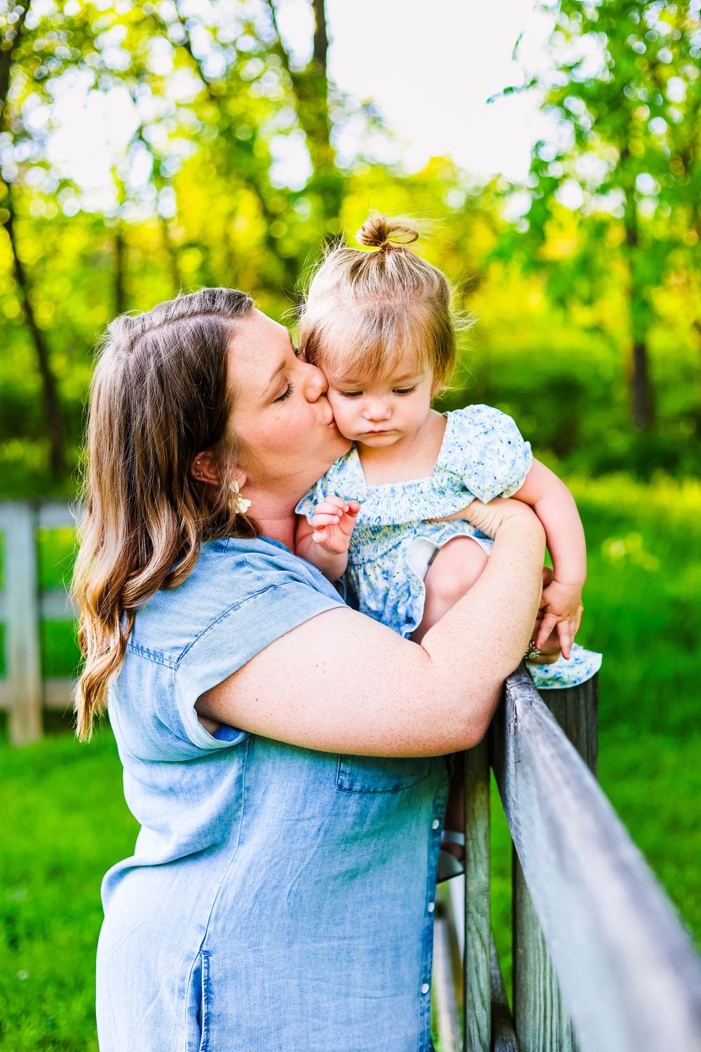 A candid/ lifestyle image of a mother kissing her baby on the cheek while sitting on a fence. Mommy and Me Session. Golden Hour.