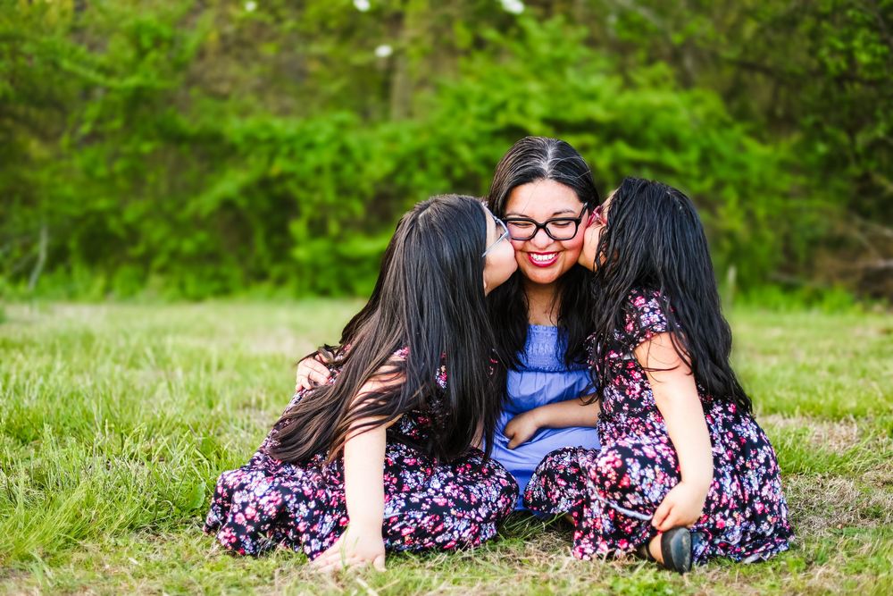 A candid image of a mother and two daughters kissing her on the cheeks sitting together in a field.