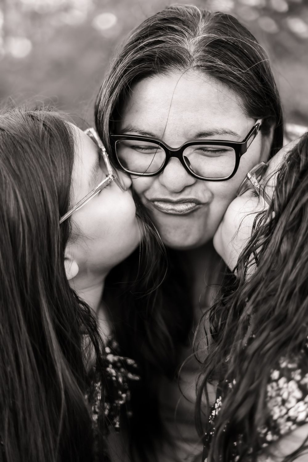 A candid black and white image of two girls kissing their mother on the cheek.