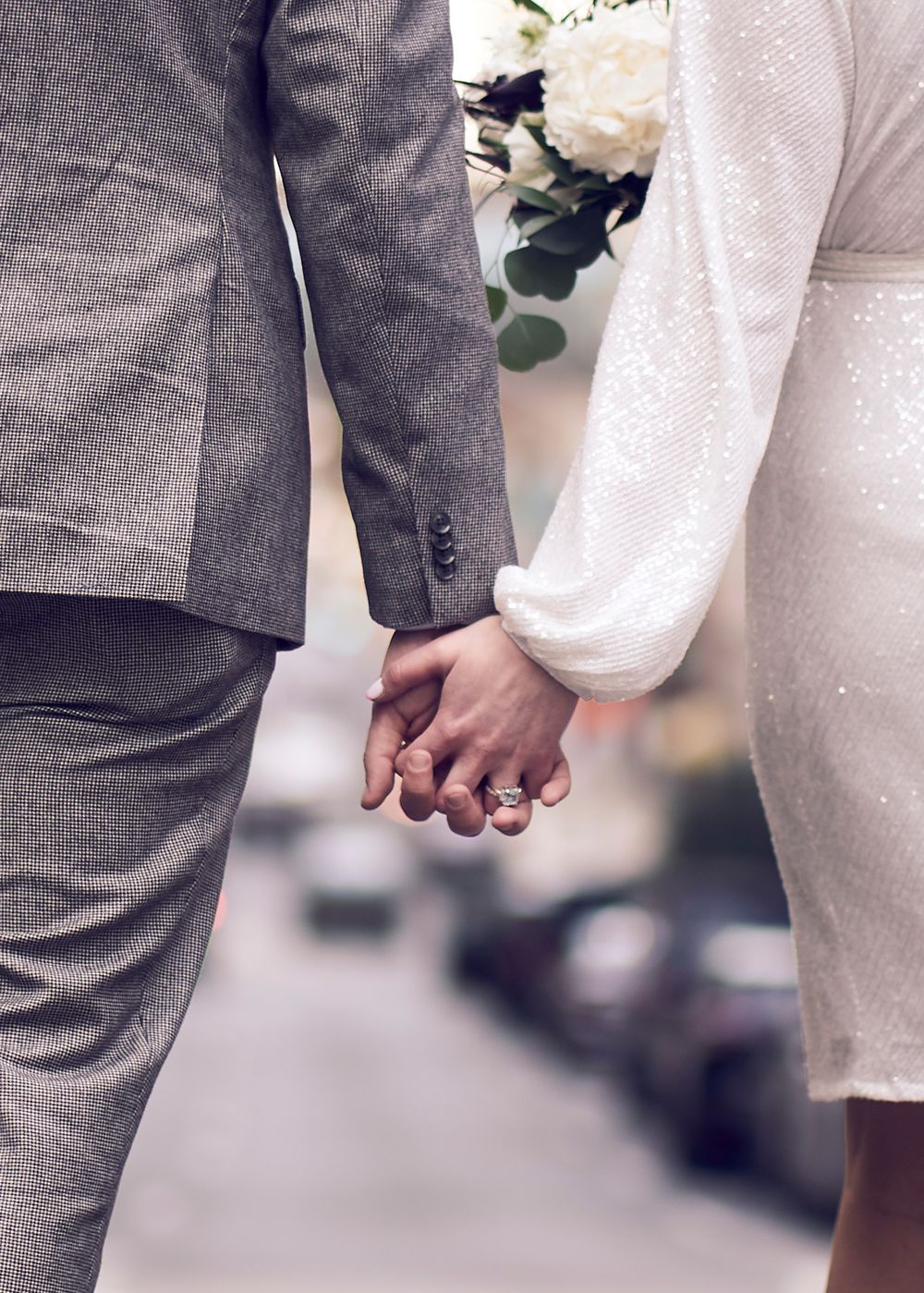 Close-up view of a newlywed couple holding hands, with bride in white dress and groom in gray checkered suit.