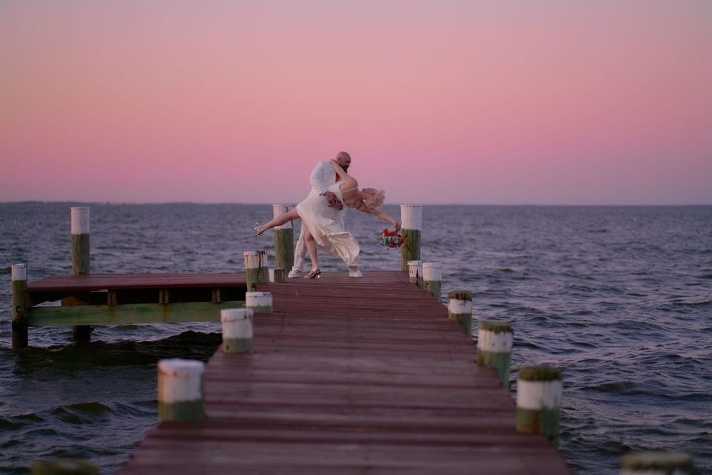 A wooden pier extends over calm waters beneath a pink and purple sunset sky at dusk.