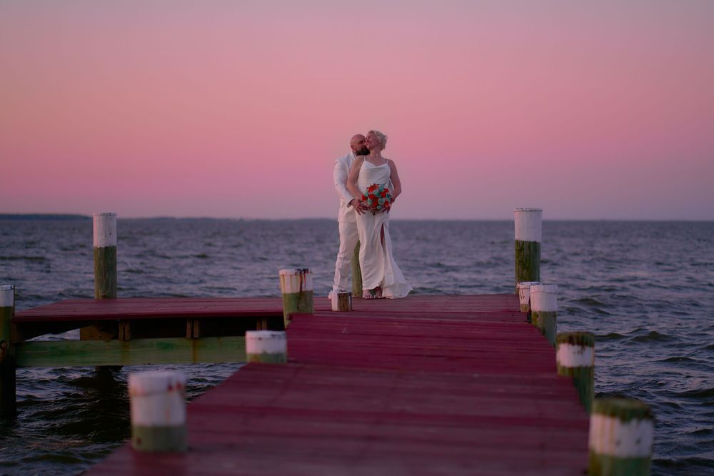 A couple in white embraces on a romantic pier with glowing lanterns at sunset over the ocean water.