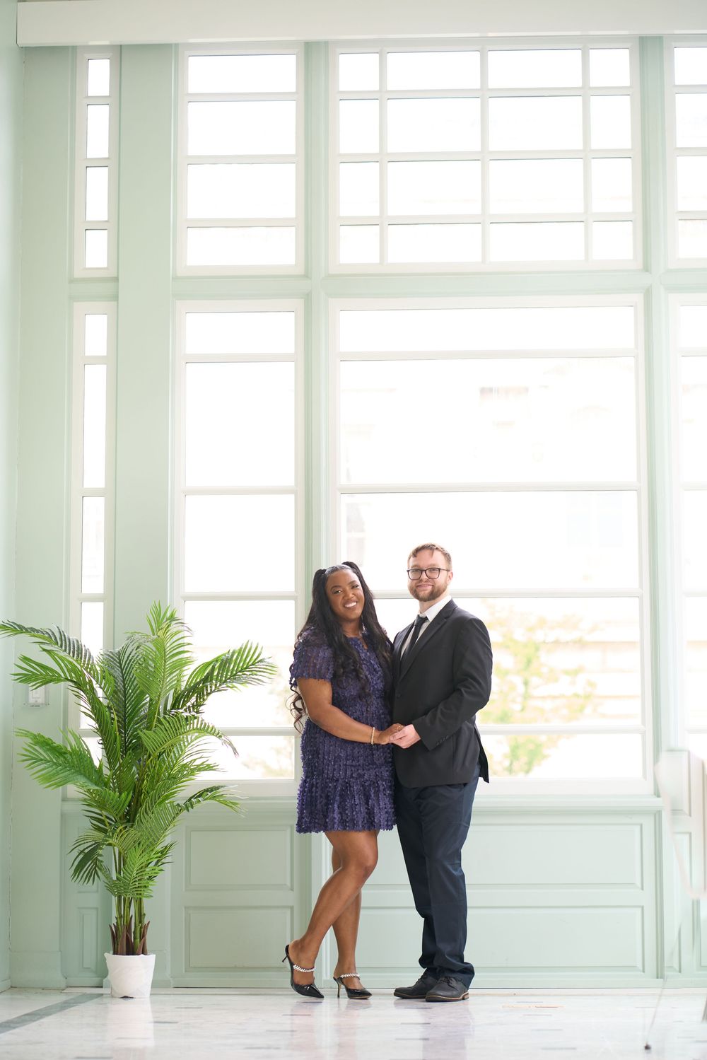 A couple shares romantic poses in a bright white room with large windows and a decorative palm plant.