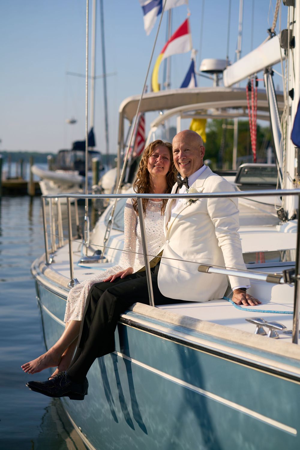 A couple in elegant evening attire sits together on a sailboat docked in a marina on a sunny day.