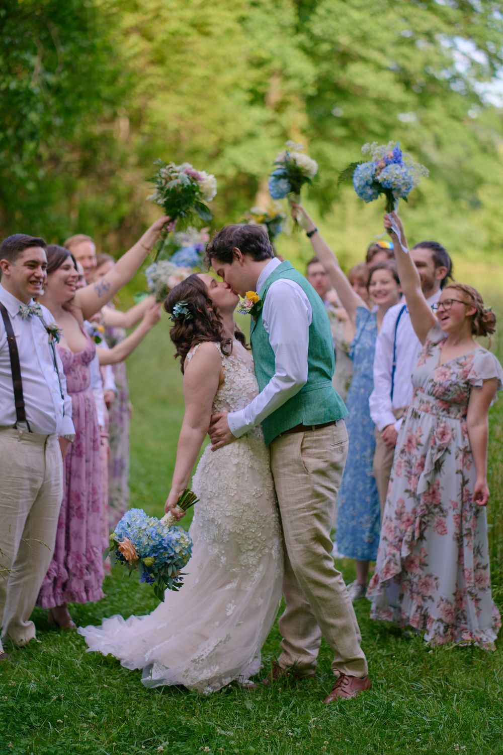 Wedding party celebrates outdoors with joyful poses in pastel floral dresses and tan suits on a green lawn.