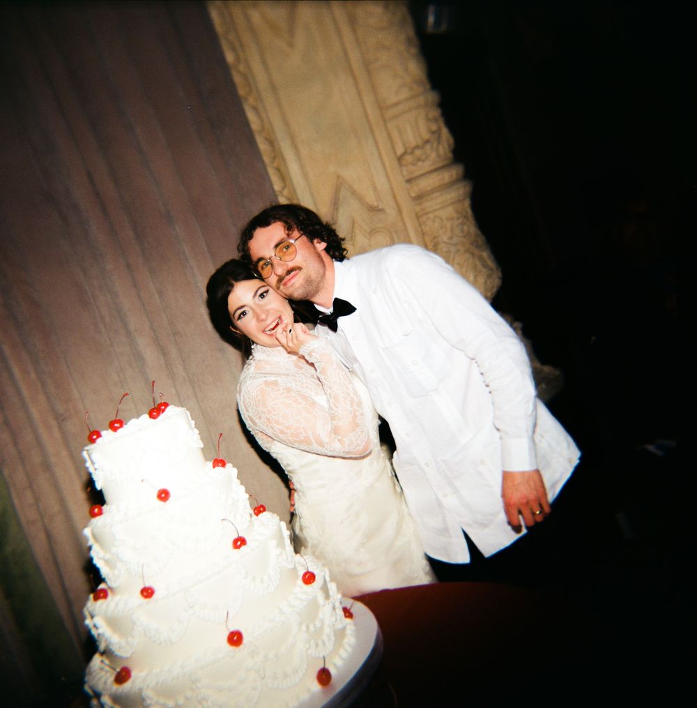 A couple shares a joyful moment beside their white wedding cake decorated with red accents at their reception.