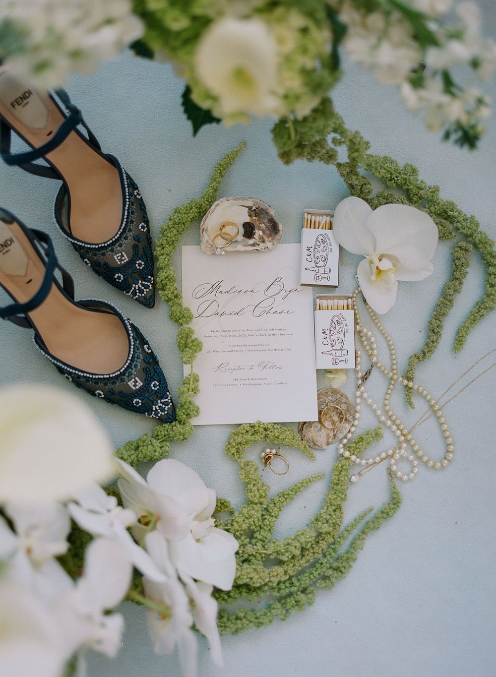 Wedding details including elegant sandals, white orchids, invitation cards, and delicate pearl necklaces arranged on a light surface.