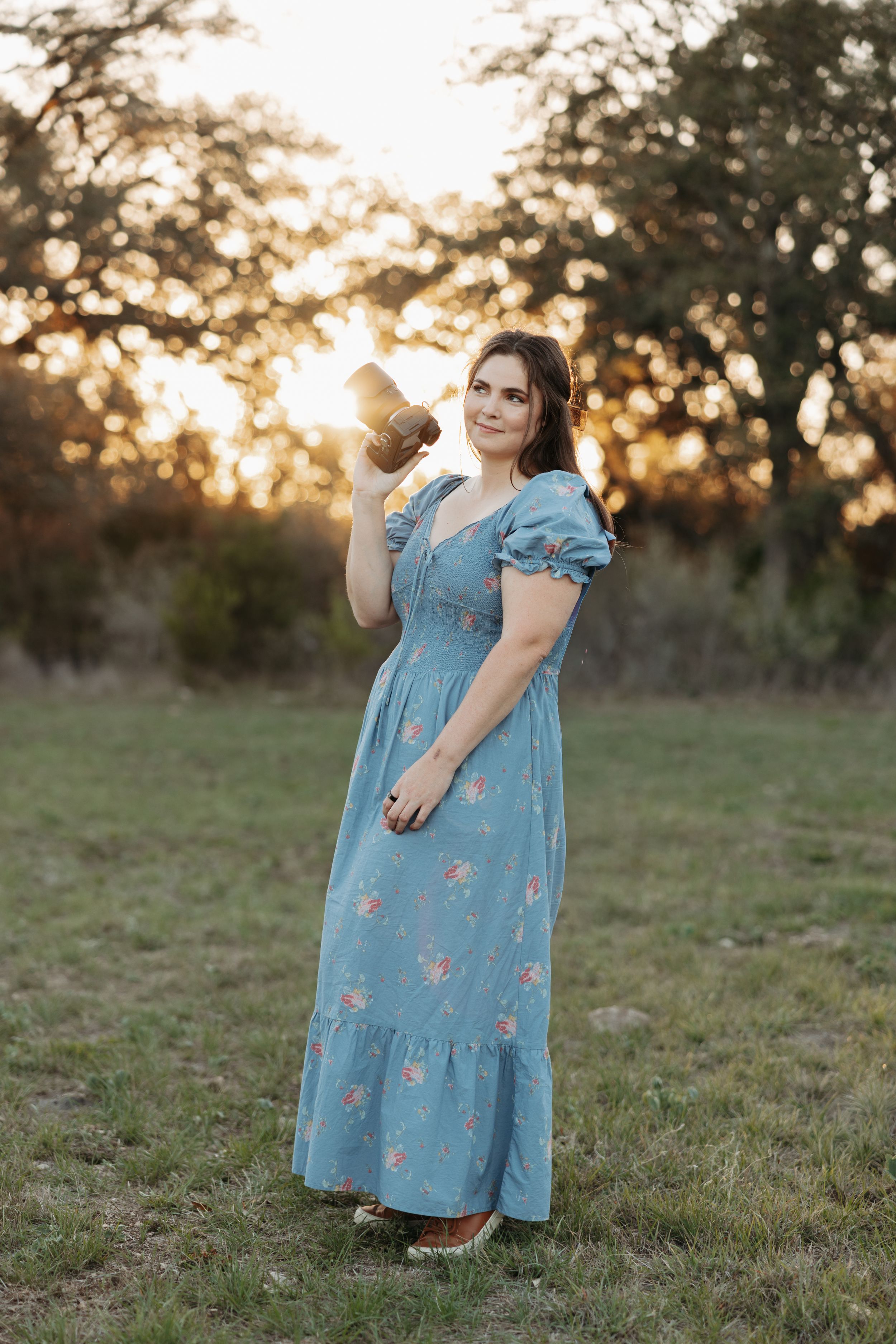 Woman in blue floral maxi dress holding a camera at golden hour in a grassy field with trees in background.