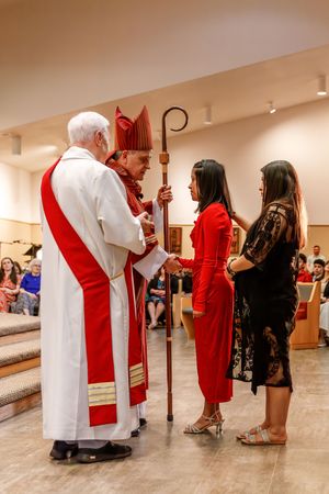 Religious ceremony with clergy in white and red vestments holding pastoral staff as attendees gather in church hall.
