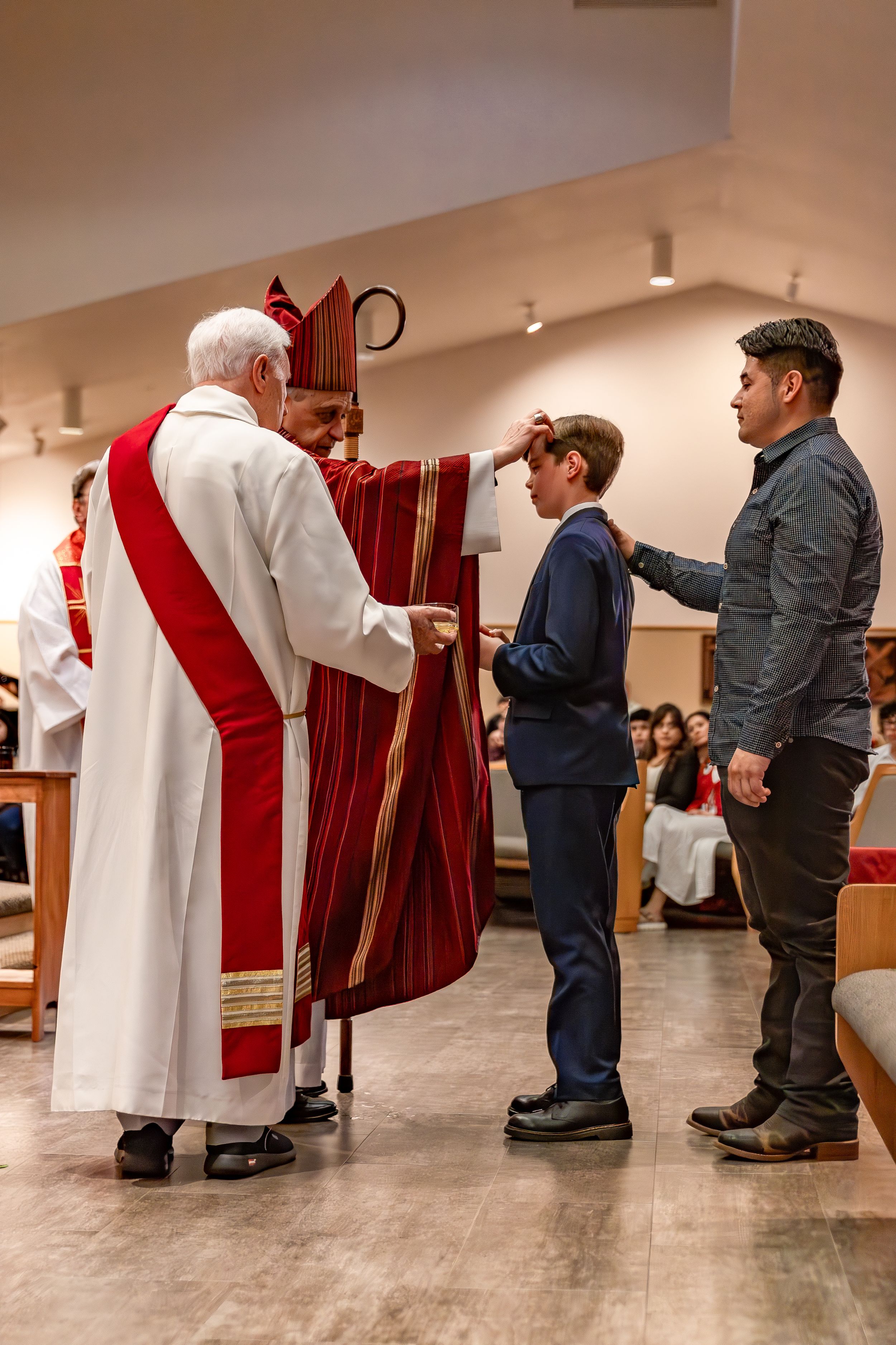 A clergyman in red and white vestments performs a religious ceremony with attendees in a church hall.