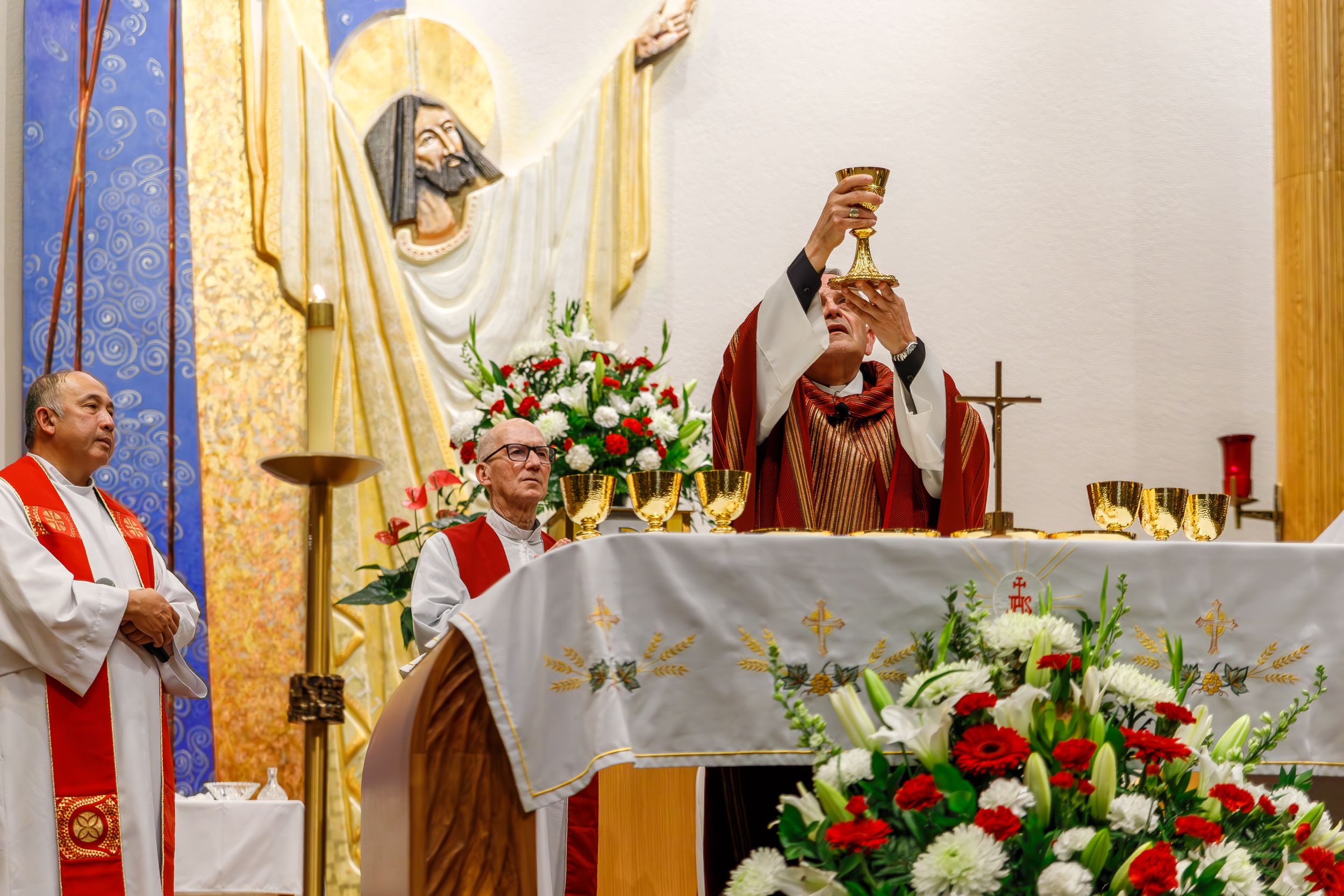 Church clergy conduct a religious ceremony at an altar decorated with red and white floral arrangements.