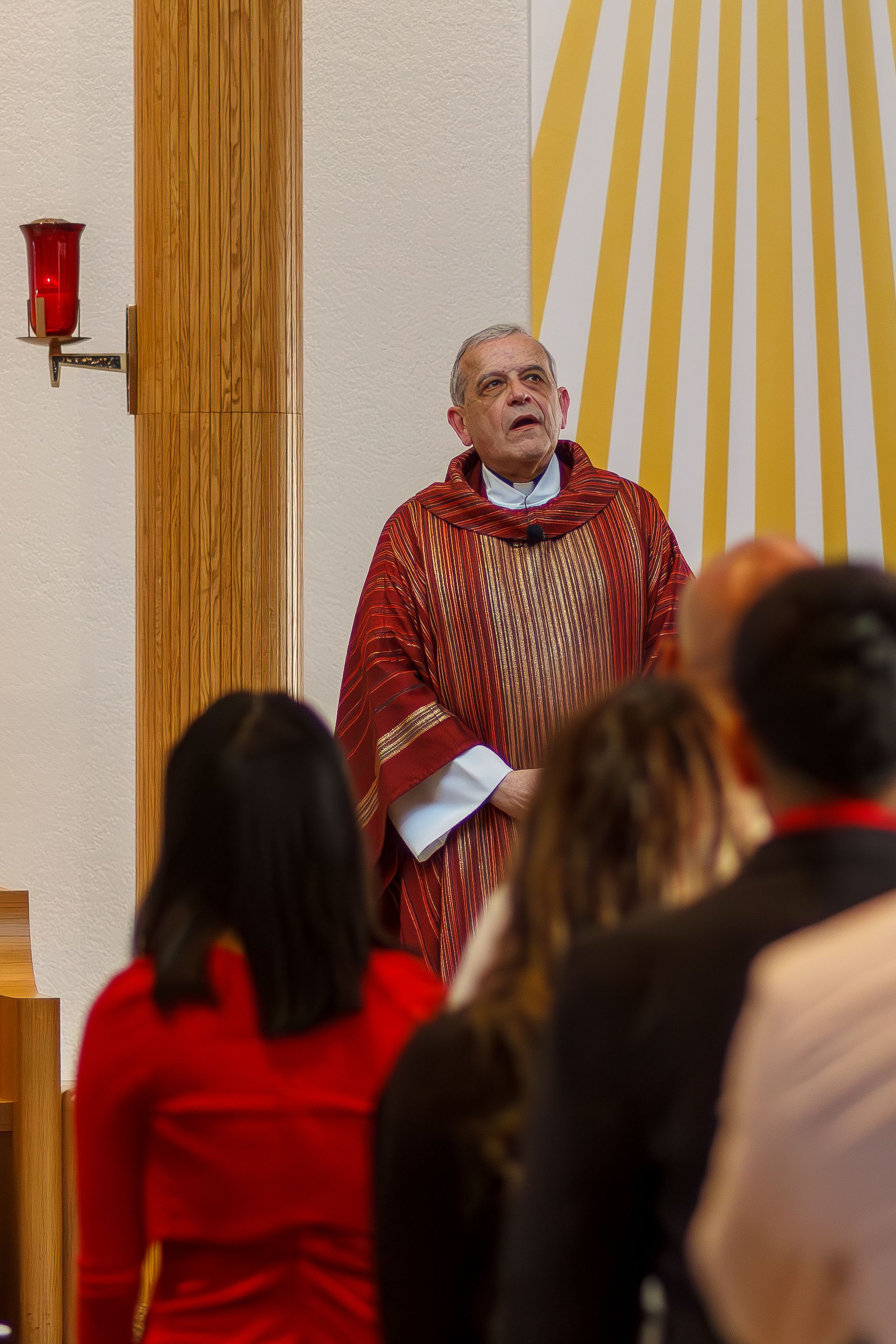 Choir members in red robes singing during a performance with wooden pillars and white walls in the background.