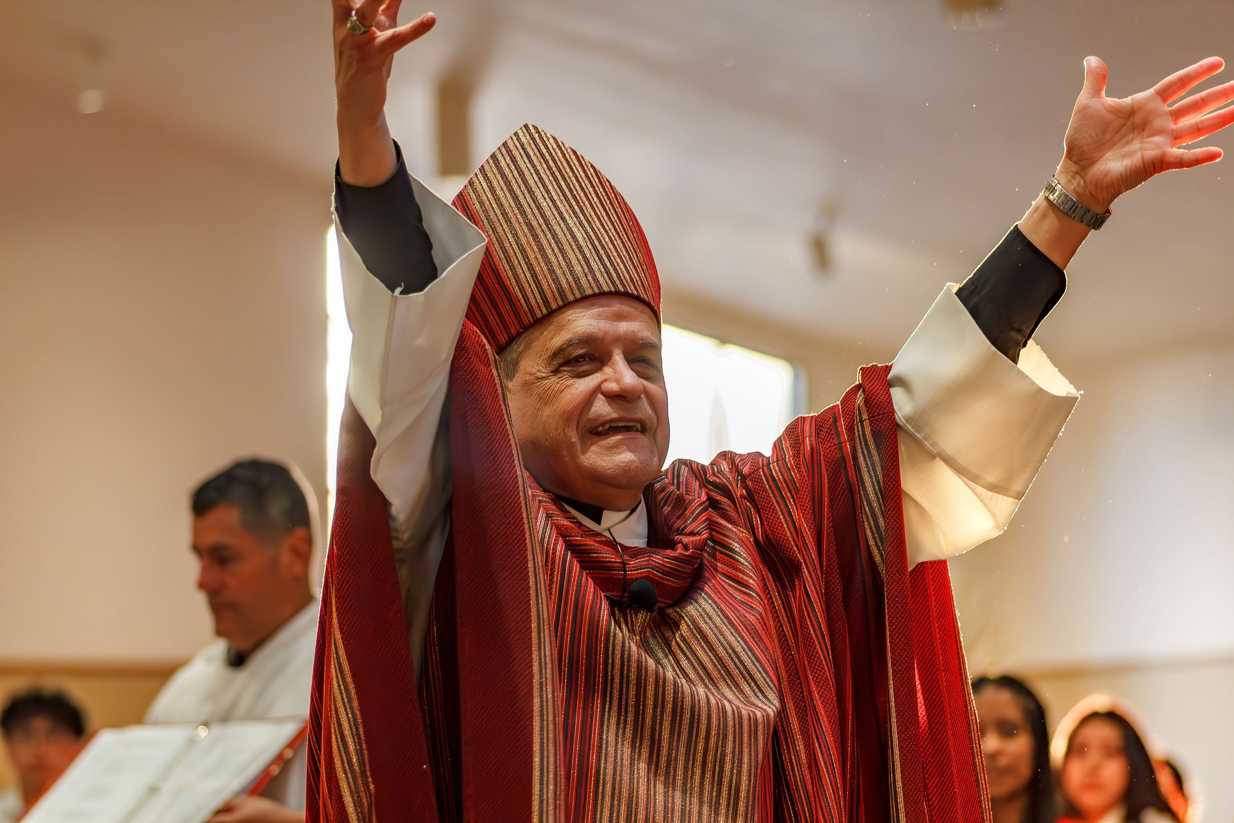 Clergy member in red ceremonial robes raising hands during worship service.