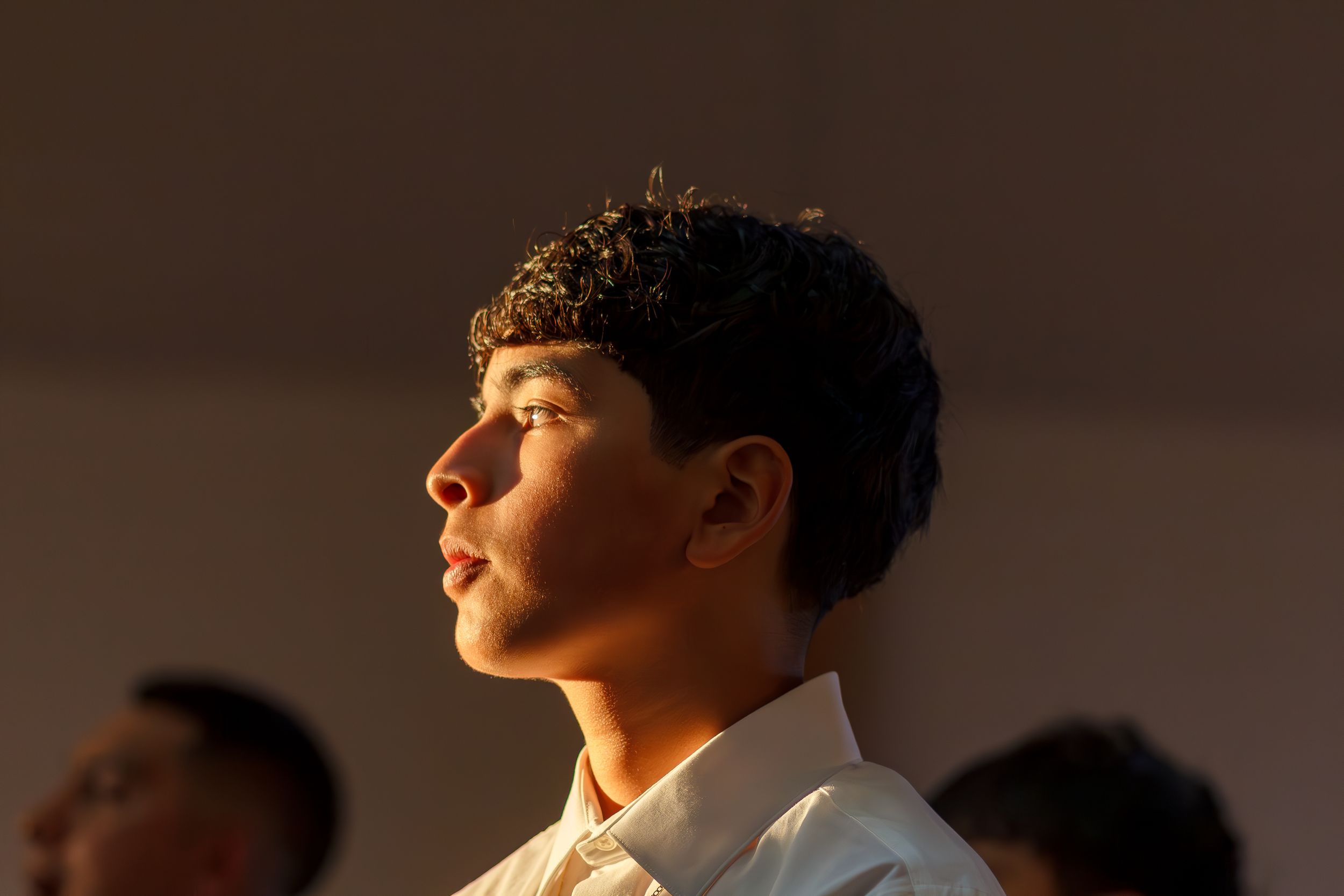 Profile view of a person in a white shirt standing in warm sunlight against a dark background.