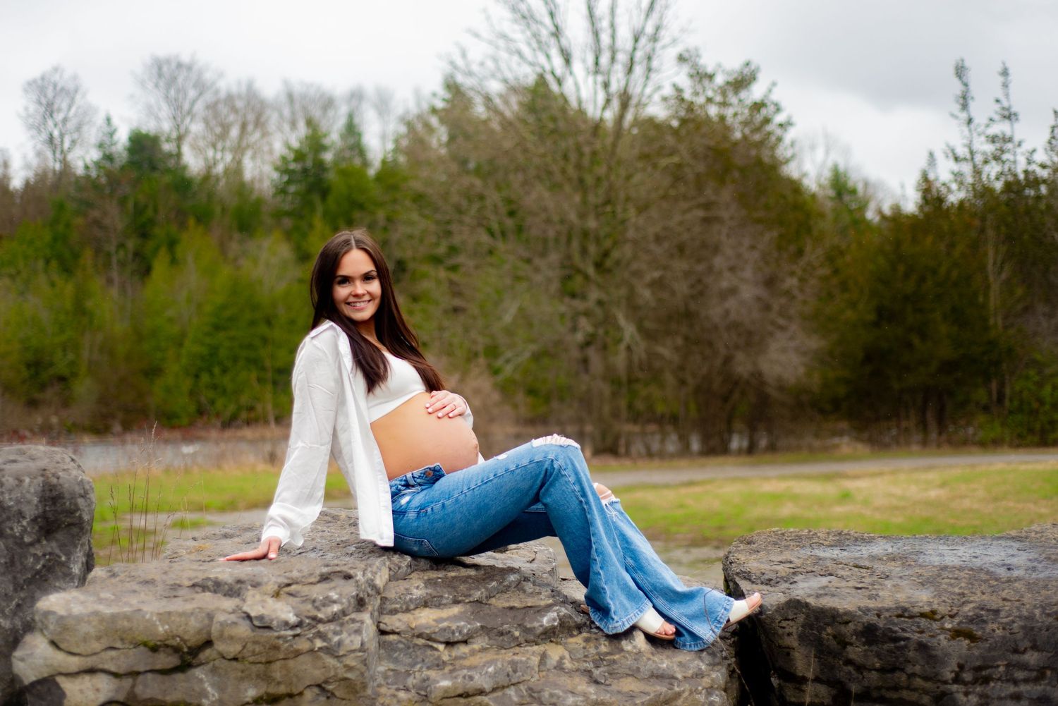 Expecting mother in casual attire relaxes on rocky outcrop during outdoor photo session.