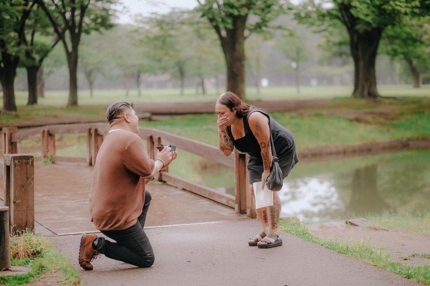 A romantic proposal moment on a wooden bridge in a misty park surrounded by green trees and a lake.