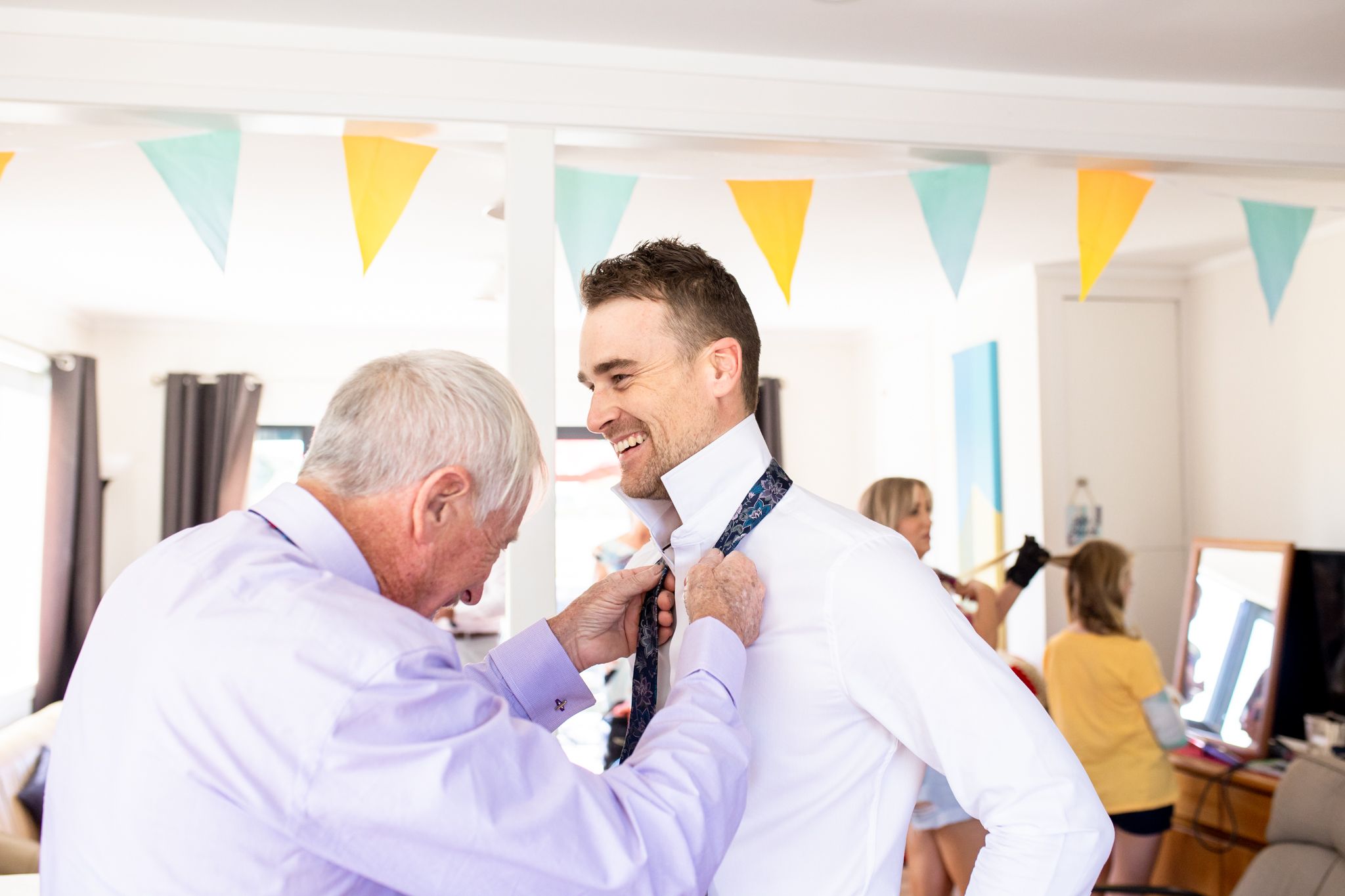 Wedding preparation scene with men adjusting formal attire under festive bunting.